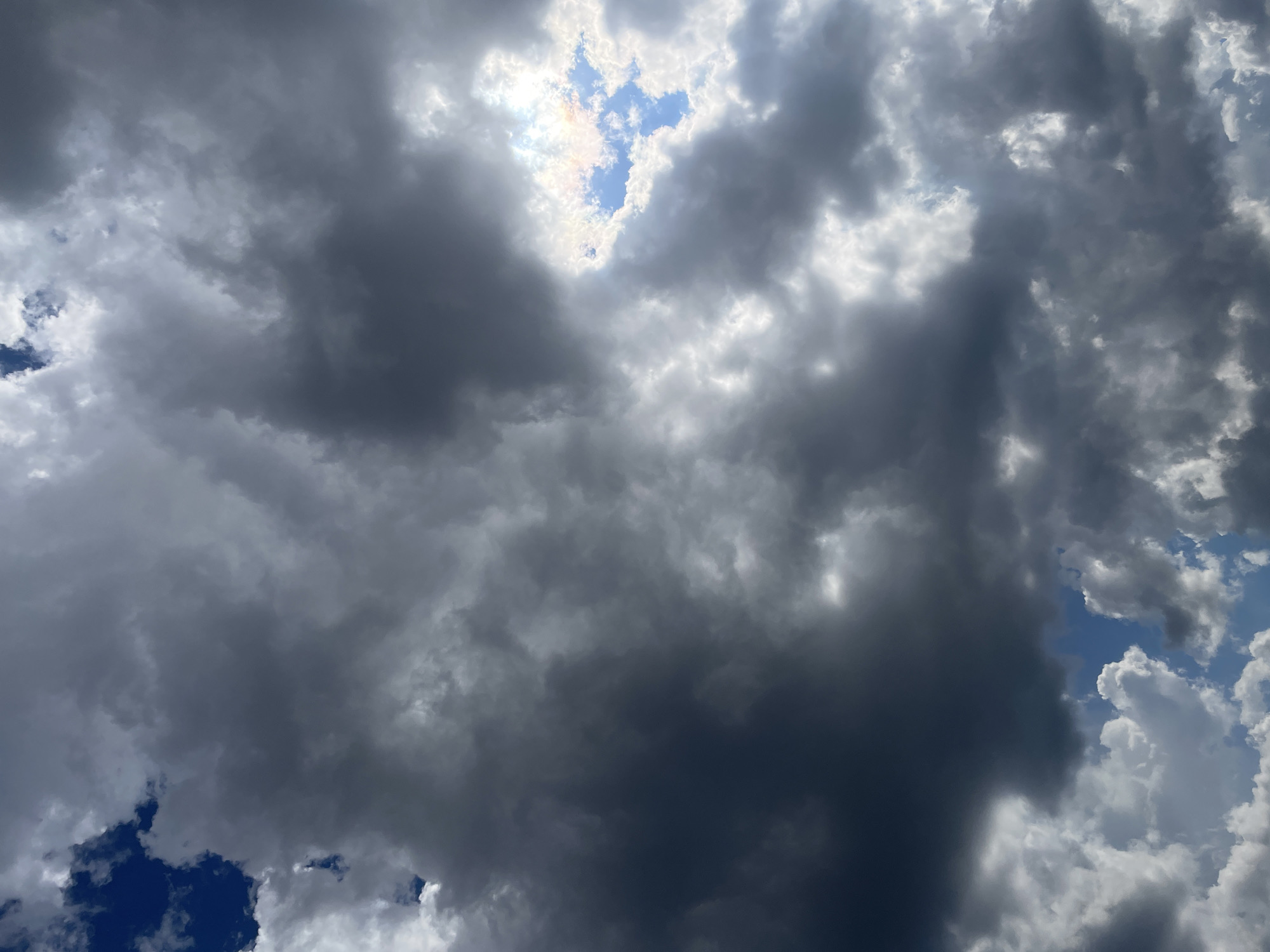 This image shows a sky with dramatic clouds. Dark, heavy clouds dominate the frame, with patches of bright, white clouds interspersed. The sun is partially visible, breaking through a gap in the clouds, creating a bright, luminous area with hints of rainbow colors. The sky is a mix of deep blues and grays, indicating a stormy or overcast day. There are no people, subjects, or objects visible in this image, only the natural atmospheric phenomenon. The location in Rostov-on-Don, Russia, is not discernible from the visual information in the image itself.