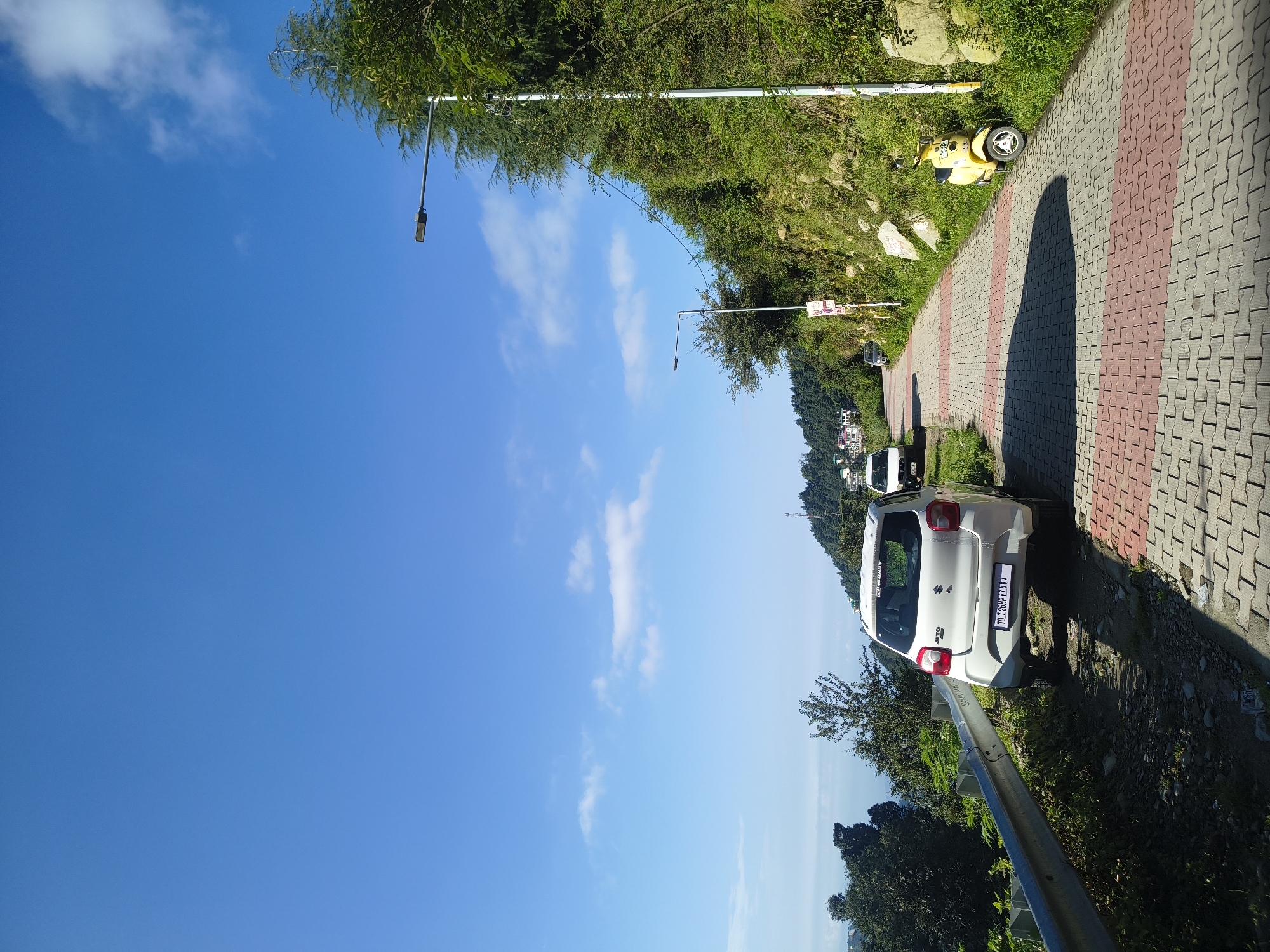 In the city of Haini, India, a white car is parked on the side of a paved road. The road is made of red and grey interlocking bricks and is bordered by a metal guardrail on the left. Lush green vegetation covers the hillside to the right of the road, with trees and bushes growing densely. A yellow scooter is parked on the right side of the paved path, near the hillside. In the distance, there are buildings visible on a hill, and a vast expanse of blue sky with scattered white clouds. The scene appears to be a scenic location, likely a tourist spot or a residential area in a hilly region.