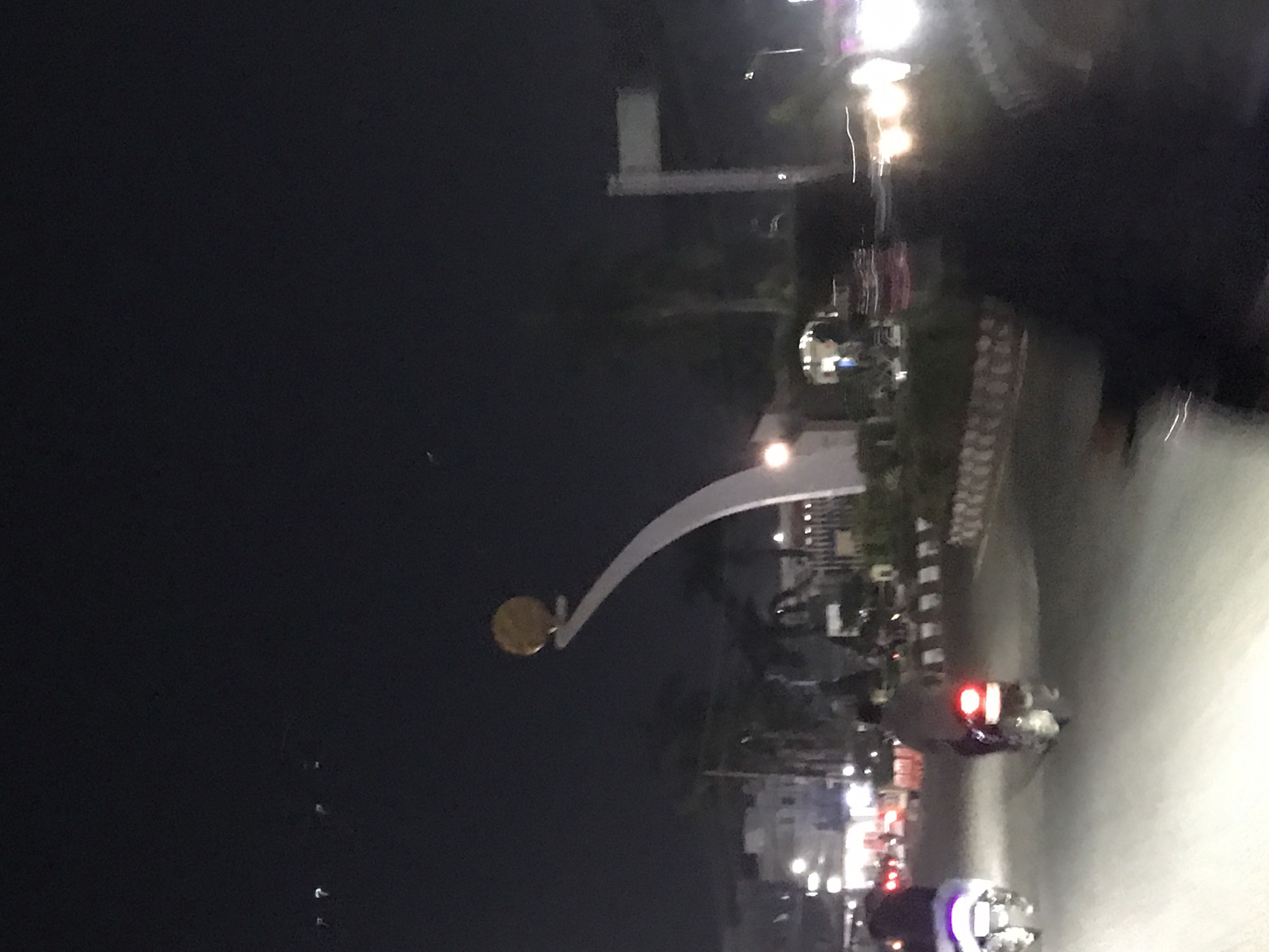 The image depicts a night scene in Lhokseumawe, Indonesia. A prominent landmark is visible: a white archway with a golden circular emblem atop a pole. Several motorcycles with illuminated headlights and taillights are present on the road. Streetlights illuminate buildings and the road itself. The environment appears to be a semi-urban road with vegetation and buildings alongside it. The photo seems to have been taken while moving, leading to motion blur, especially on the right side.