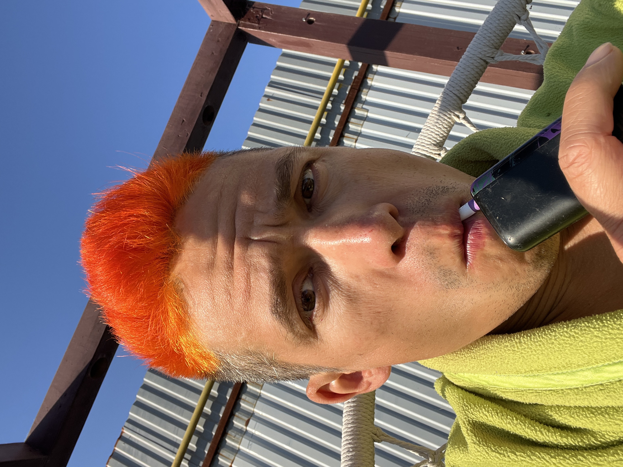 A close-up, rotated image shows a person with orange hair and a slight frown. They are holding a black electronic cigarette to their lips. The person wears a lime green robe, and behind them, there's a structure made of wooden beams and corrugated metal. The blue sky is visible in the background. The location is Chaikovsky, Russia.