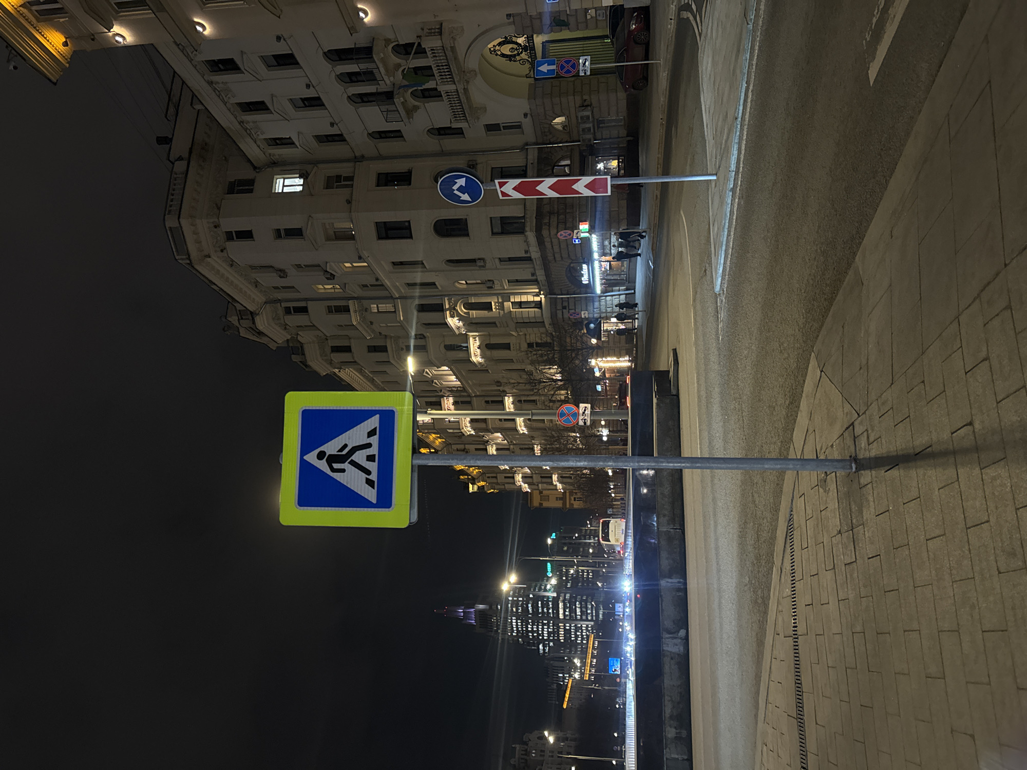 The image captures a nighttime urban scene in Moscow, Russia, specifically looking eastward from the vicinity of the Bolshoy Ustinsky Bridge towards the Kotelnicheskaya Embankment Building.
In the foreground, a grey-tiled sidewalk extends from the lower left. A bright, illuminated square blue pedestrian crossing sign, featuring a white walking figure on a blue triangle with a yellow border, is centrally positioned on a grey pole. The pole's shadow extends across the sidewalk. To the right, an asphalt road surface begins. Further right, another grey pole holds a circular blue sign with two white arrows (up and down) and, beneath it, a red and white chevron hazard sign.
The midground features a multi-lane roadway, likely a bridge segment, with visible vehicle headlights indicating ongoing traffic. A low concrete barrier separates this road from the foreground.
The background on the right is dominated by a multi-story, light-colored historical building featuring numerous windows, some illuminated from within, balconies, and ornate architectural details. Several pedestrians are visible on the sidewalk in front of this building. A ground-level establishment, partially visible, displays "О'Рыба" (O'Fish). A red passenger vehicle is parked on the far right.
To the left of the center, across the roadway and in the distance, the prominent Stalinist-era Kotelnicheskaya Embankment Building stands tall, characterized by its numerous illuminated windows and a distinctive purple-lit spire. The sky is dark, indicating night, with various streetlights and building illuminations providing ambient light to the scene.