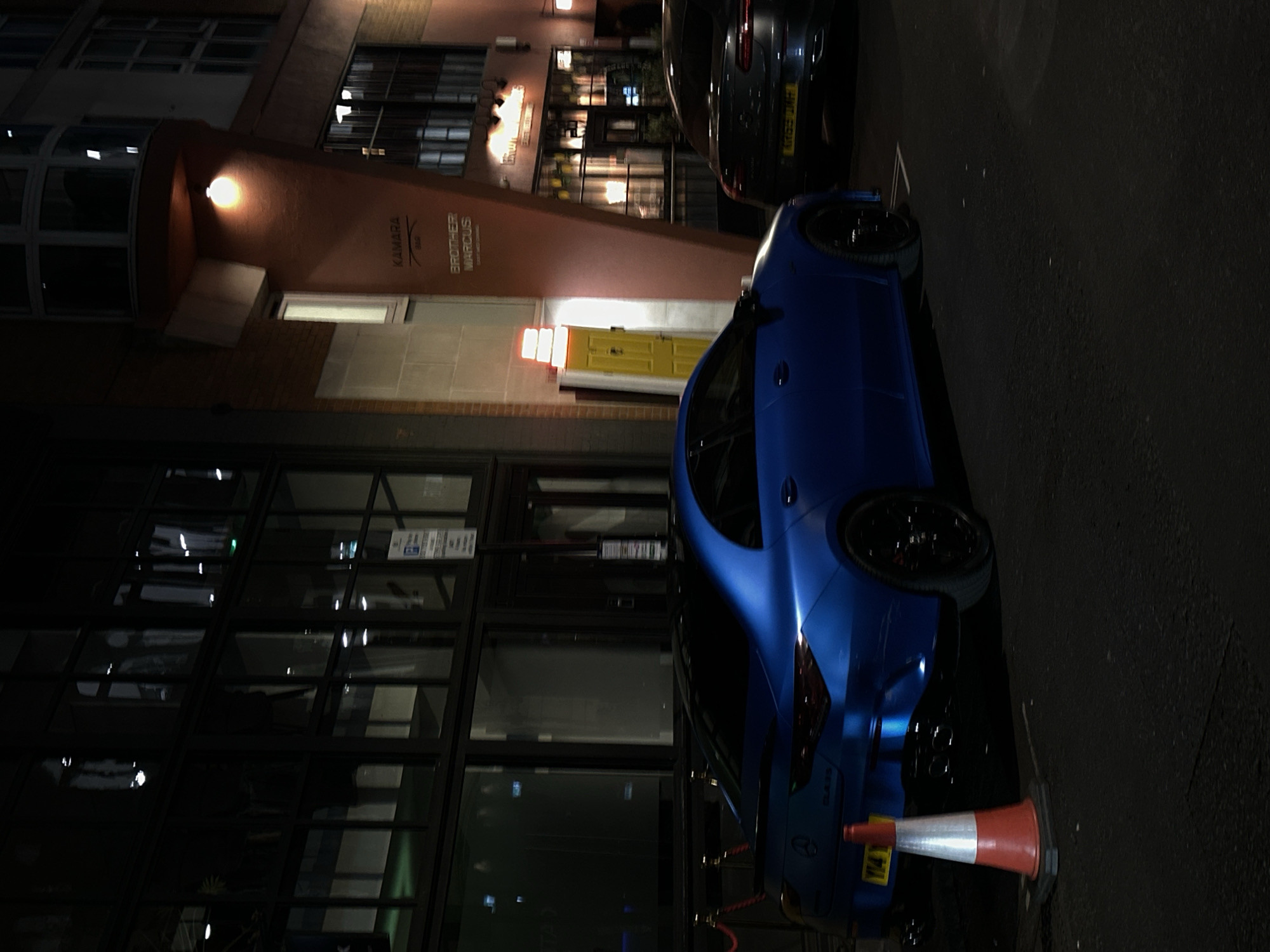 The image shows a blue Mercedes-Benz AMG parked on a street in London, United Kingdom. A traffic cone is positioned behind the vehicle, close to the bumper. A black car is parked in front of the blue one. Across the street, there's a building with illuminated signs reading "KAMARA" and "BROTHER MARCUS". There is a yellow door in the background. The building has large windows and appears to be a business or restaurant. It's nighttime, and the street is illuminated by streetlights and building lights. The surrounding environment includes urban buildings and infrastructure.