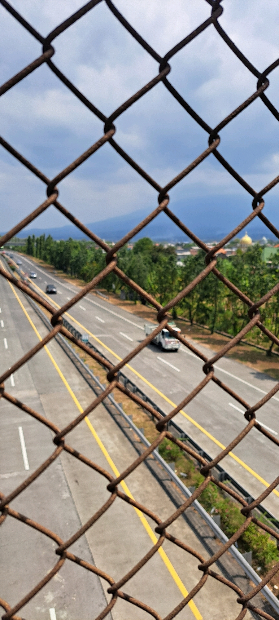 This is a vertical, eye-level shot taken through a rusty chain-link fence, looking down at a multi-lane highway in Kebon Waris, Indonesia. The fence is in the foreground, with its diamond-shaped mesh prominent and out of focus. The highway stretches into the distance, with clear lane markings, including double yellow lines. Several vehicles are visible on the highway, including cars and a white truck. The highway is flanked by green trees and vegetation. In the background, a city skyline with buildings and a golden-domed structure is visible, with rolling hills or mountains in the far distance under a cloudy sky. The overall scene depicts a typical urban highway environment in Indonesia.