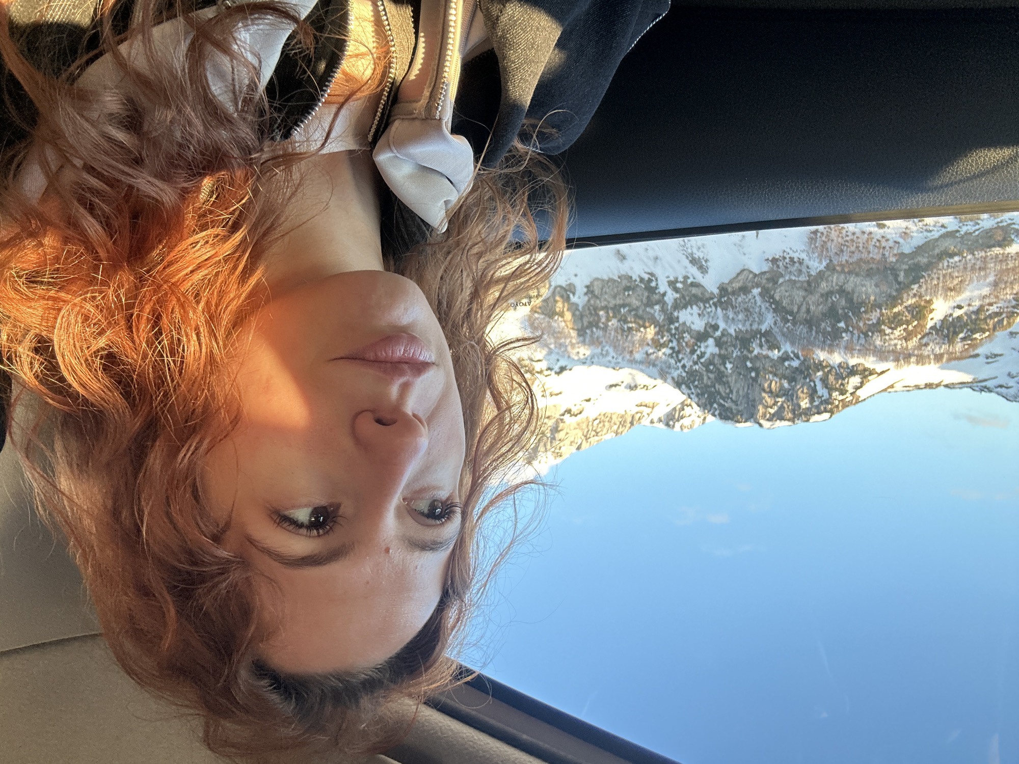 A woman with reddish-brown curly hair is pictured from a low angle. She is fair-skinned with neutral lip color and brown eyes. She is wearing a jacket with white trim and a zipper. Through the window above her, snow-covered mountains are visible under a clear blue sky. The location is not discernible from the image. There are no apparent actions or interactions.