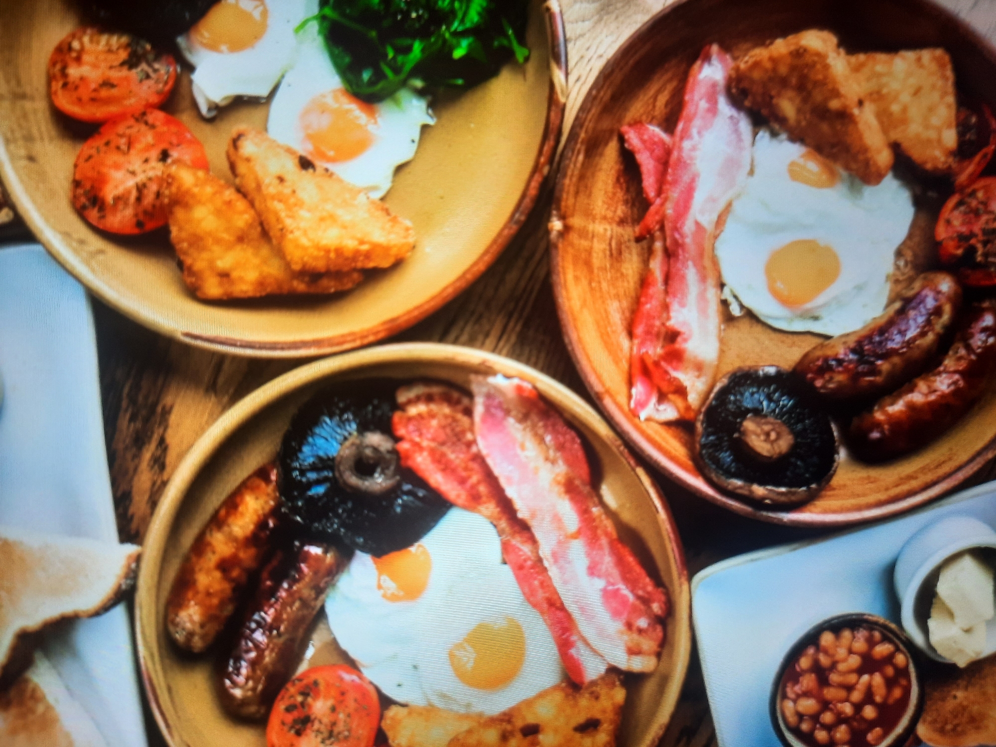 The image displays a close-up aerial view of three plates of food, likely a full English breakfast, arranged on a wooden surface. The food is presented in a rustic, slightly disheveled manner.
The left plate contains two sausages, a grilled mushroom, grilled tomato halves, two fried eggs, and strips of bacon. The middle plate, slightly to the right and higher, features two fried eggs, grilled tomato halves, hash browns, and a pile of wilted green leafy vegetables, possibly spinach. The right plate, lower down and to the right, shows two sausages, a grilled mushroom, strips of bacon, hash browns, and a fried egg. In the lower left corner, a small white dish contains a dollop of what appears to be butter or cream cheese, and a small bowl is filled with baked beans. There are also pieces of toasted bread visible, one in the upper left corner and another below the butter dish. The lighting is warm and diffused. There are no people or any indication of Rosenheim, Germany.