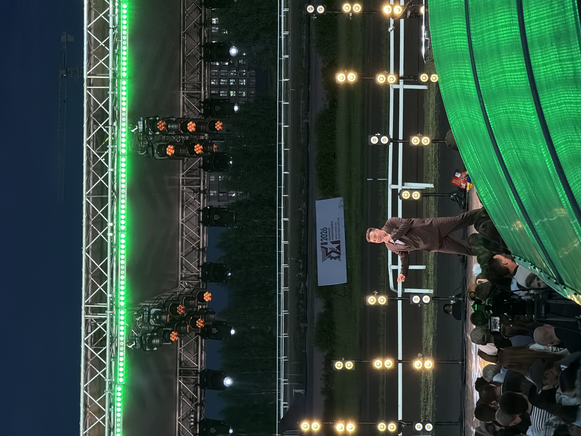The image depicts an outdoor evening event, possibly a concert or festival, at a location that appears to be a racecourse or an open field with stadium-like lighting. In the foreground, a crowd of people is gathered, with some holding up phones. A person is visible in the center of the frame, on a stage, wearing a dark suit and holding a microphone, appearing to be performing or speaking to the audience. The stage is illuminated by a series of bright lights, including green linear lights overhead and multiple smaller yellow lights on stands behind the performer. In the background, there's a large white banner with text and a graphic, and beyond that, trees and buildings under a dark sky. A large construction crane is visible in the upper part of the image. The overall scene suggests a lively event taking place at dusk or after dark.