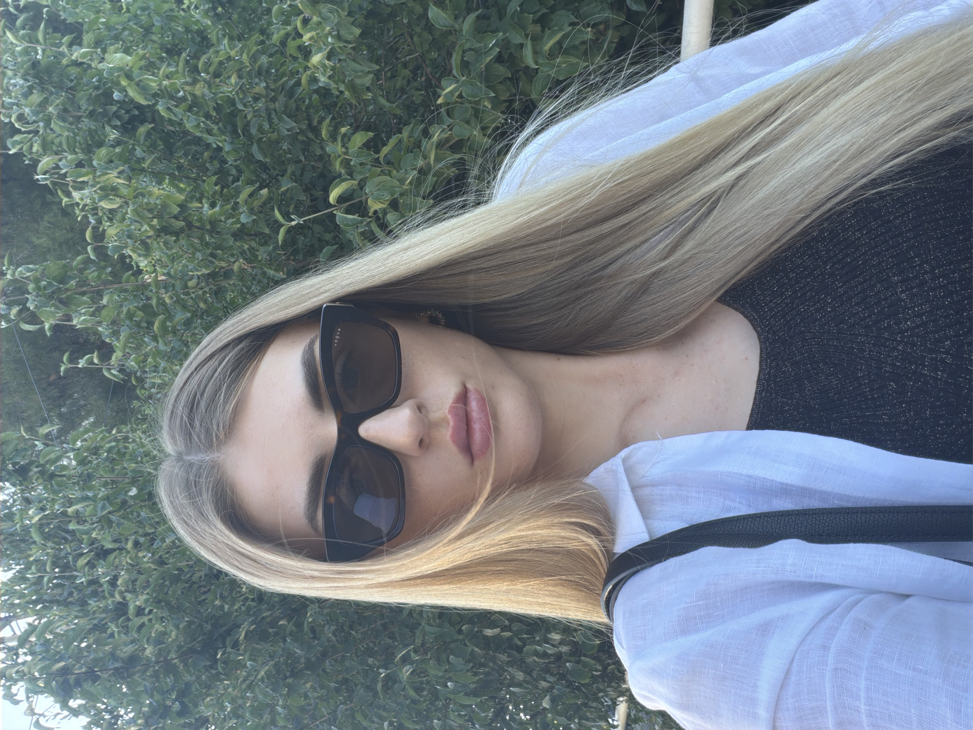 This is a close-up, vertically oriented, selfie-style photograph of a young woman. She has long, straight blonde hair that is parted in the middle and cascades around her face. She is wearing large, dark-rimmed sunglasses with a tortoiseshell pattern. Her lips are painted a subtle pink. She is wearing a white, linen-like shirt with the sleeves rolled up and a black, sparkly top underneath. A dark strap, likely from a bag, is visible across her shoulder.
The background is dominated by lush green foliage, suggesting an outdoor setting, possibly in a garden or near trees. The lighting appears natural and bright, indicating it's likely daytime, with the sun casting a warm glow on her hair and skin. There are no overt signs of a specific event or activity taking place; the image captures a moment of leisure or casual personal documentation. The overall mood is relaxed and stylish. There is a small, white pole-like object visible at the top right of the image, which could be part of a fence or railing. No text is visible in the image.