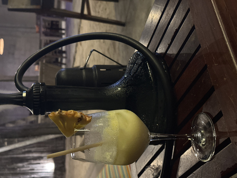 A sideways view showcases a frosty yellow cocktail garnished with a pineapple wedge and straw, sitting on a wooden slatted surface. Behind the drink, a black metal structure with a looped handle and cylindrical component dominates the frame. The backdrop is blurred, suggesting an outdoor setting with faint hints of architectural elements and vegetation. The scene is lit softly, casting a cool hue.
