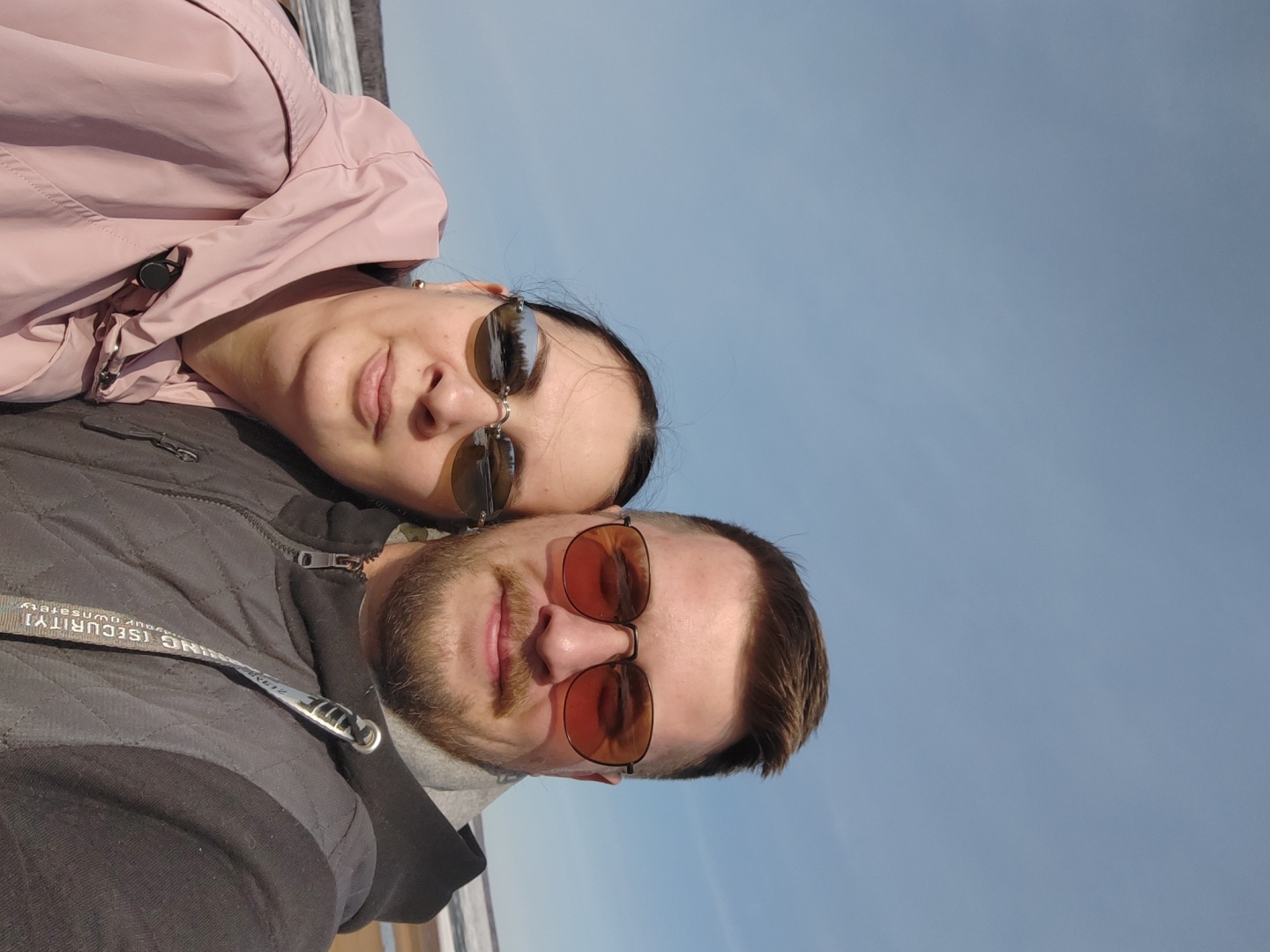 A couple is posing for a selfie outdoors. The woman, wearing a pink jacket and sunglasses, is lying on top of the man. The man, with a beard and sunglasses, is wearing a gray jacket. The background shows a blue sky. The photograph was taken in the city of Chaikovsky, Russia.