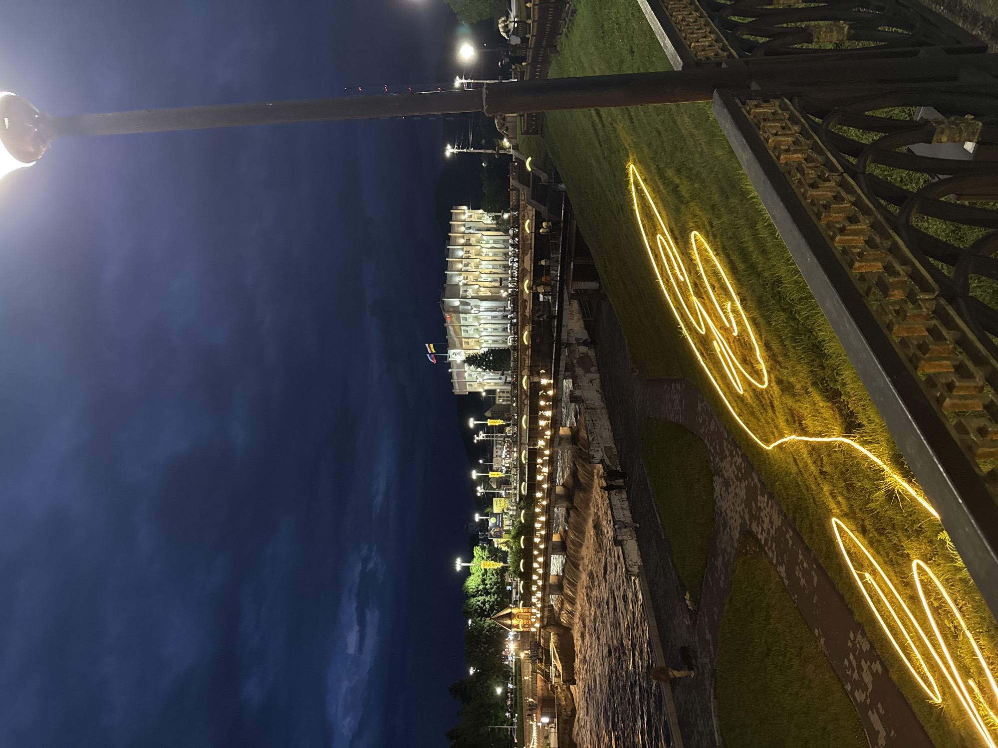 The image captures a nighttime view of a waterfront promenade in Vladikavkaz, Russia. The scene is illuminated by a combination of streetlights and decorative yellow neon outlines that trace organic shapes on the grassy areas. In the background, a multi-story building with numerous lit windows suggests urban activity. The sky is a deep blue, indicating it's after sunset, and the presence of clouds hints at a possibly cool or overcast evening.
Along the promenade, there's evidence of pedestrian activity, with a few silhouetted figures visible. The waterfront itself appears to be a river or canal, with its surface reflecting the artificial lights. A notable feature is an ornate black metal fence in the foreground, which frames the view and adds to the sense of a curated public space. The presence of trees and manicured lawns suggests a park-like setting designed for public enjoyment. The overall atmosphere is tranquil and somewhat festive, owing to the lighting decorations. No discernible text is visible within the image.