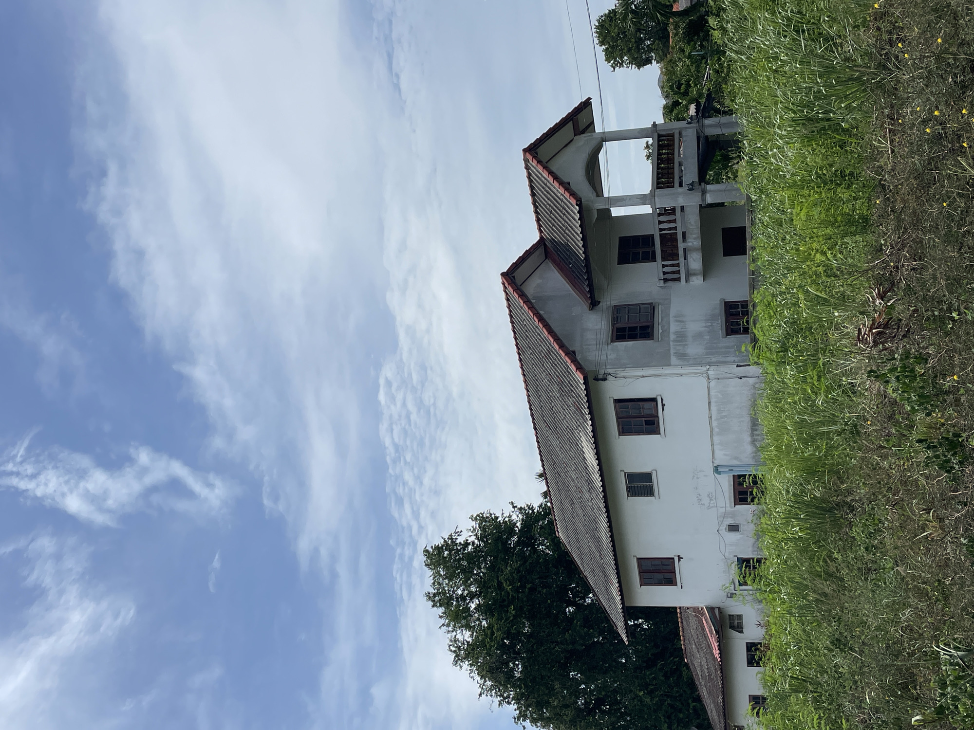 A two-story white house with a red-tiled roof is visible in the image. The house appears to be old and somewhat weathered. It is situated amidst lush green vegetation, with a large tree to the left of the house. The sky above is blue with scattered white clouds. The scene depicts a residential area in the city of Muang Pattaya, Thailand. There are no people or visible actions in the image.