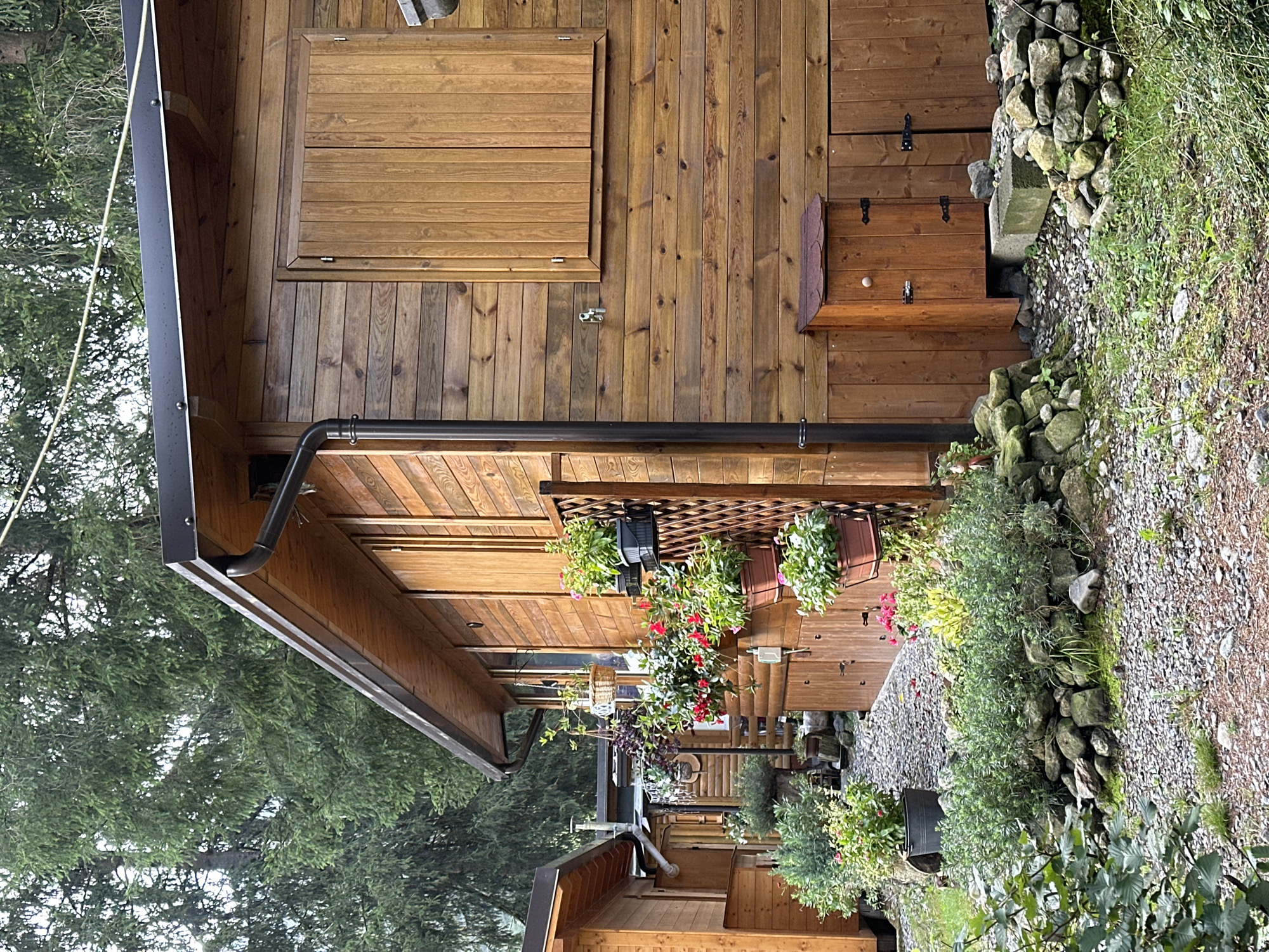 The image depicts a row of wooden cabins situated in Borno, Italy, nestled amongst lush greenery. The cabins, constructed from natural wood, feature pitched roofs with dark gutters and downspouts. One cabin in the foreground has a closed wooden shutter on its window and a small wooden storage unit with a hinged door at its base. The area around the cabins is landscaped with gravel paths and various potted plants, including flowering bushes and hanging baskets, adding a touch of vibrant color to the rustic setting. The surrounding environment is dominated by tall evergreen trees, suggesting a mountainous or forested location. The lighting indicates it is daytime, possibly overcast due to the soft light and absence of strong shadows. No people are visible in the image.