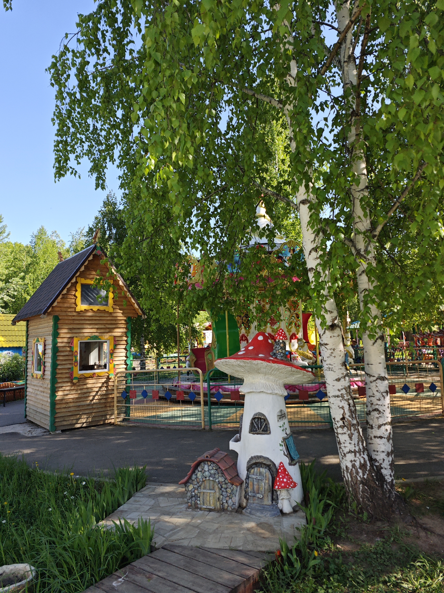 The image depicts a scene at a children's park in Chaikovsky, Russia. There is a small wooden playhouse, resembling a log cabin with a black roof and yellow window trim. Nearby stands a mushroom-shaped structure with a red-spotted cap, designed as a fairy tale house, complete with a small door. A birch tree with white bark is positioned next to the mushroom house. In the background, a colorful carousel can be seen, suggesting a recreational or amusement park setting. The ground is paved, with some grass and vegetation.