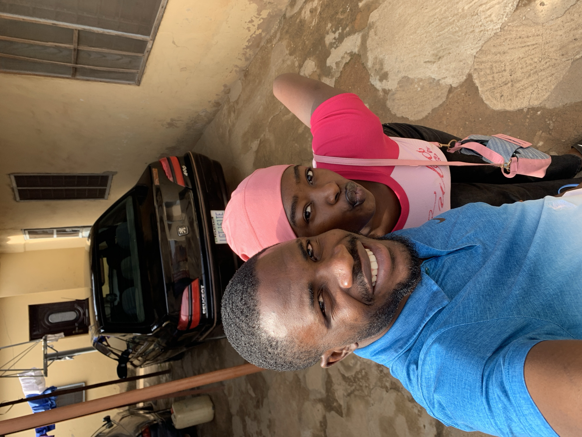 The image shows two individuals, a man and a woman, posing closely together for a selfie. The man, in the foreground, is wearing a blue shirt and has a beard. The woman, positioned behind him, is wearing a pink head covering and a pink and white top with a handbag slung across her shoulder.
Behind them, a black Peugeot 406 car is visible, parked against a yellow wall. Above the car, there is a window with a visible metal grill. In the far background, a door is partially visible. The setting appears to be an outdoor courtyard with concrete ground, possibly located in Rafin Par, Nigeria, based on the license plate of the car.