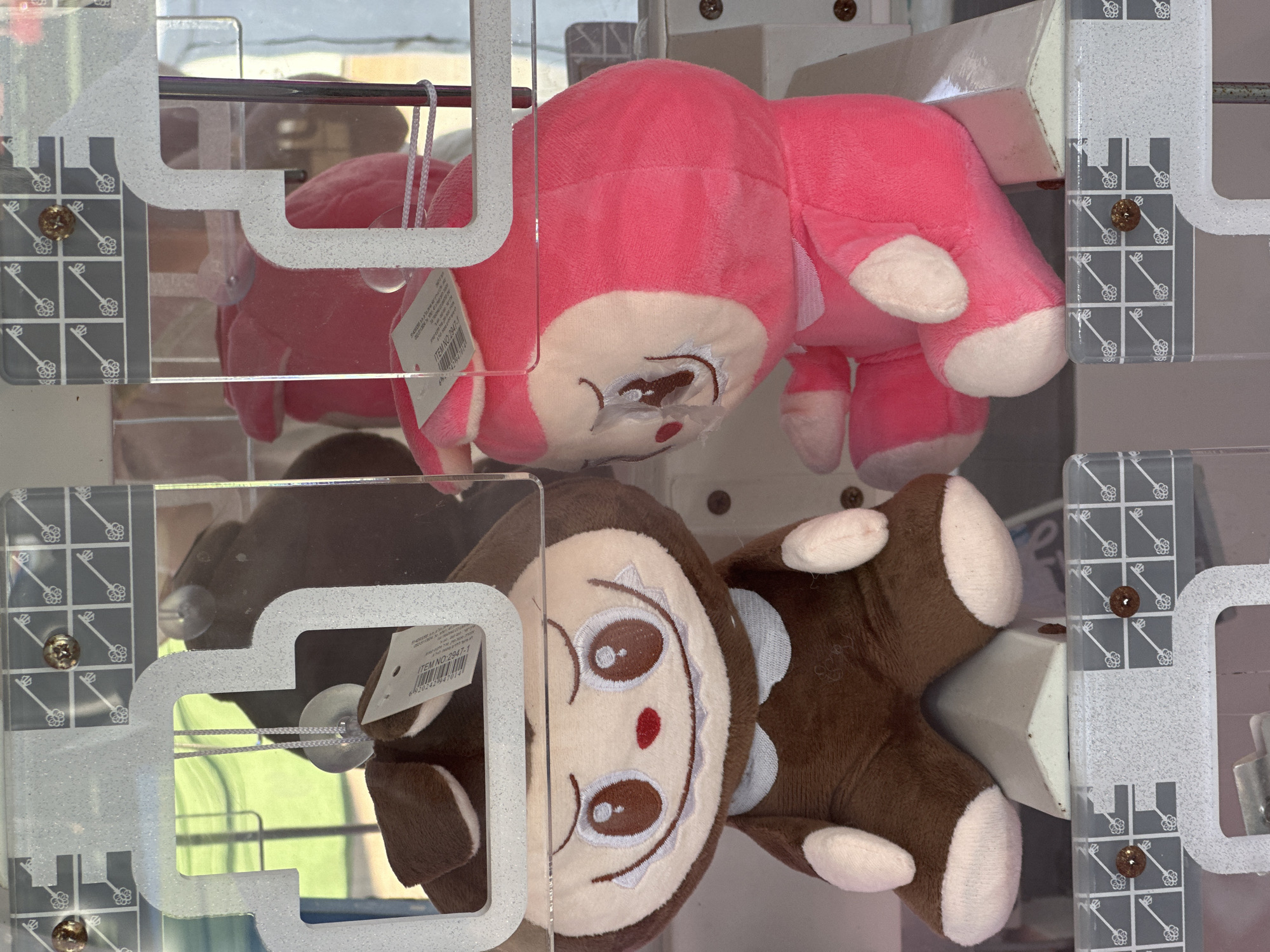 This image shows two plush toys inside a claw machine. The toy on the left is brown with a white collar and has a wide smile and large eyes. The toy on the right is pink with a white collar and also has large eyes and a smiling expression. Both toys appear to be sitting upright. The background shows a brightly lit interior with the typical clear plastic and metal framework of a claw machine. The machine is located at a "Yellow" store in Hod HaSharon, Israel, as indicated by text on a label visible in the image.