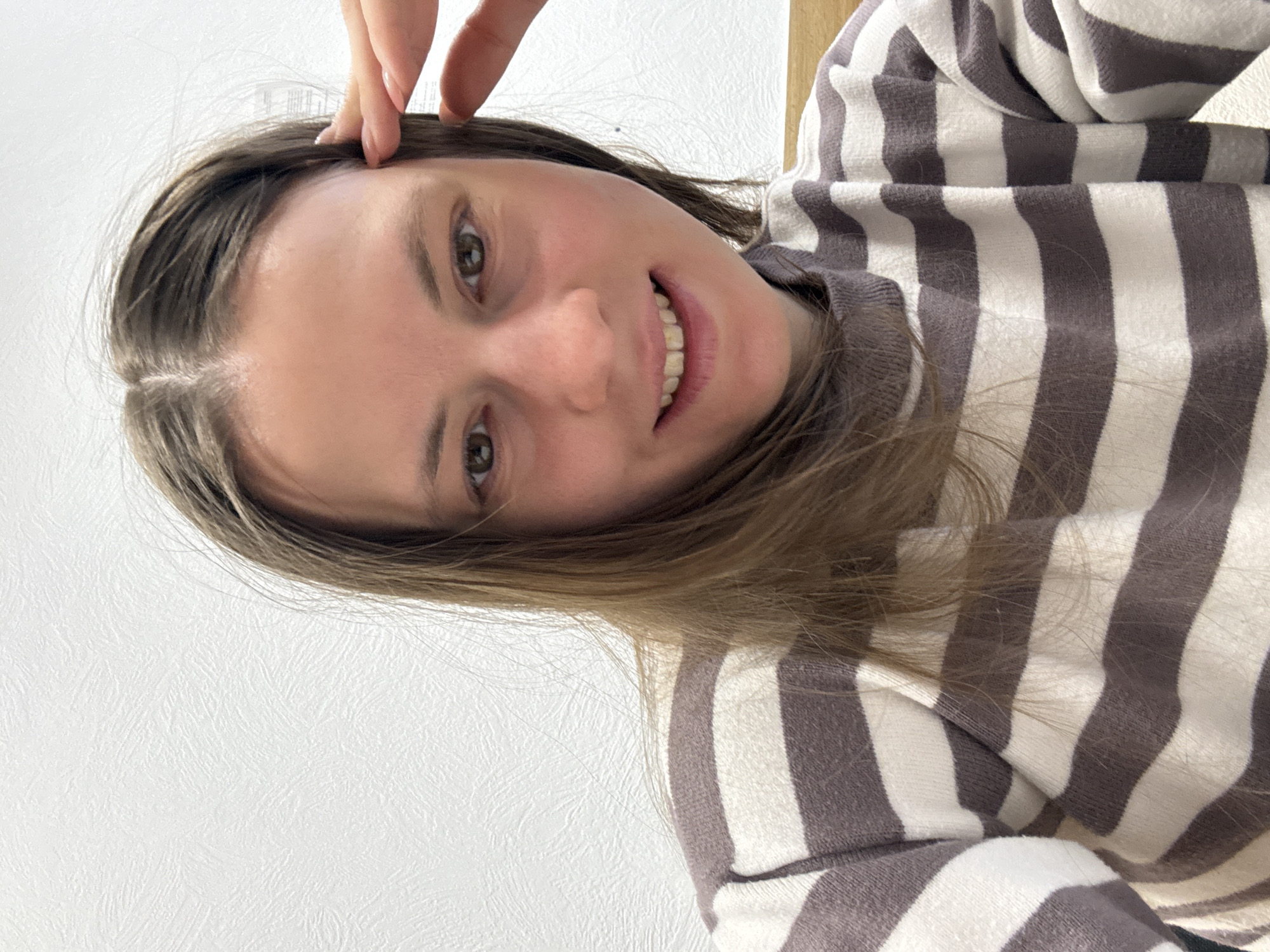 A woman with light brown hair and fair skin smiles at the camera, touching her forehead with her fingers. She wears a striped sweater. The background is a plain white wall. The location in the city of Chaikovsky, Russia, is not evident from the image.