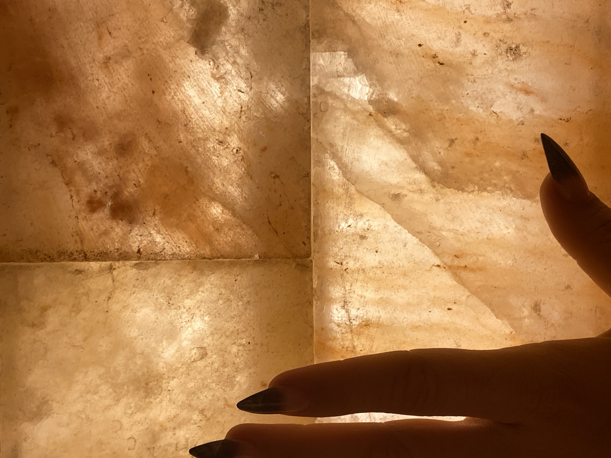 The image shows a close-up of a hand with dark, pointed fingernails resting against a backlit, textured surface that appears to be made of salt blocks. The surface is a warm, amber color with variations in tone and visible crystalline structures. The hand is partially obscuring the surface, with the fingers and thumb angled as if being held up to the light. The lighting creates a soft glow and casts subtle shadows. The scene is not identifiable as being located in Votkinsk, Russia, or any other specific city. There are no discernible elements in the image that would indicate a location.