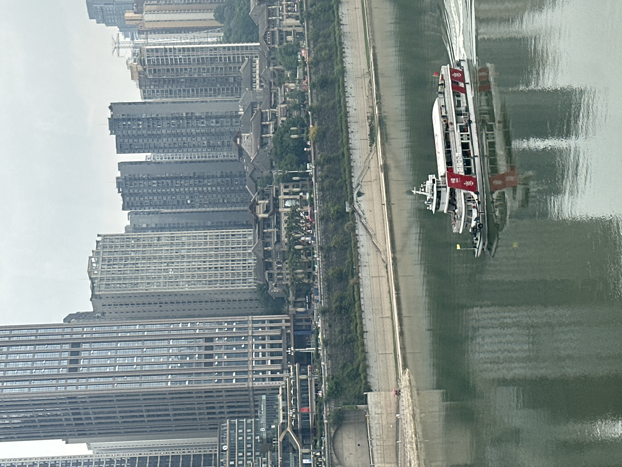 The image depicts an urban scene in Yuzhong District, China. A passenger ferry with red flags moves along a river. The riverbank features a concrete embankment. Adjacent to the embankment is a green strip with trees. The foreground shows modern high-rise buildings with glass facades. In the background are older residential buildings with tiled roofs. The sky is overcast.