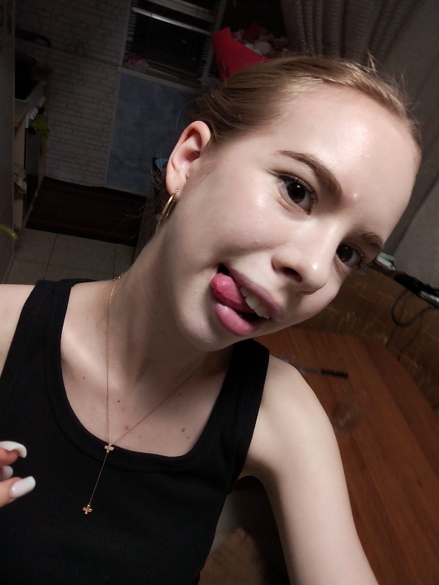 This is a close-up selfie of a young woman with light skin and blonde hair pulled back. She is wearing a black tank top and a delicate gold necklace with two cross pendants. Her right ear is visible, adorned with a gold hoop earring. She is sticking her tongue out playfully, with her mouth open and teeth slightly visible. Her eyes are dark brown and she has well-defined eyebrows. Her skin appears smooth and slightly shiny, possibly due to makeup or lighting. The background is blurry and suggests an indoor setting, with hints of a brick wall, a blue rug, and wooden furniture. The lighting is somewhat dim, with a warm tone. The overall impression is one of a casual and playful moment.