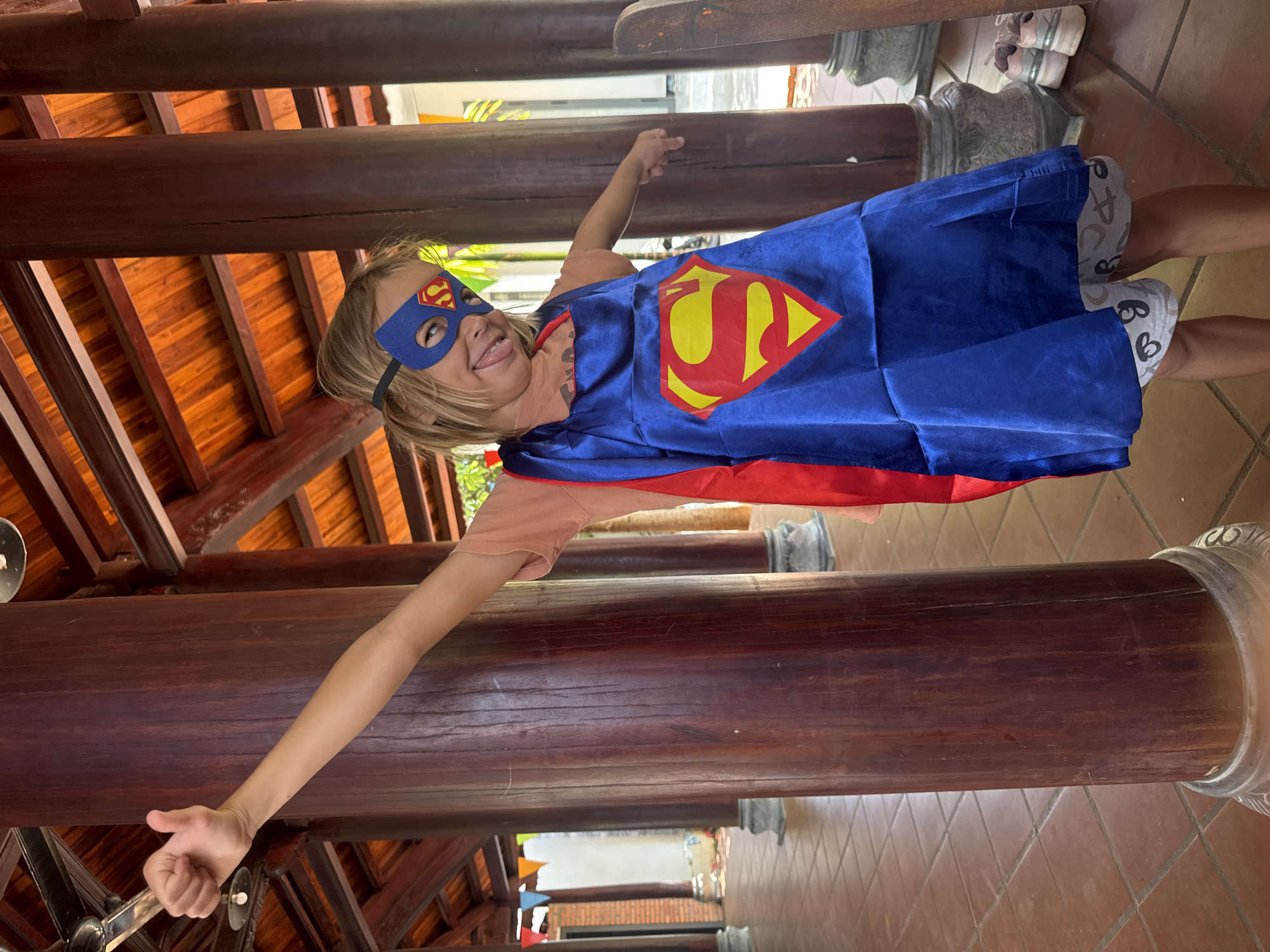 A young child in a blue Superman cape and mask strikes a superhero pose in front of an outdoor structure made of dark wood beams in Mũi Né, Vietnam. The child has blonde hair and is wearing a peach-colored shirt. The cape has a red and yellow Superman logo on the back. The structure appears to be a pergola or covered walkway with a tiled roof. The ground is tiled. In the background, there are glimpses of foliage and buildings.