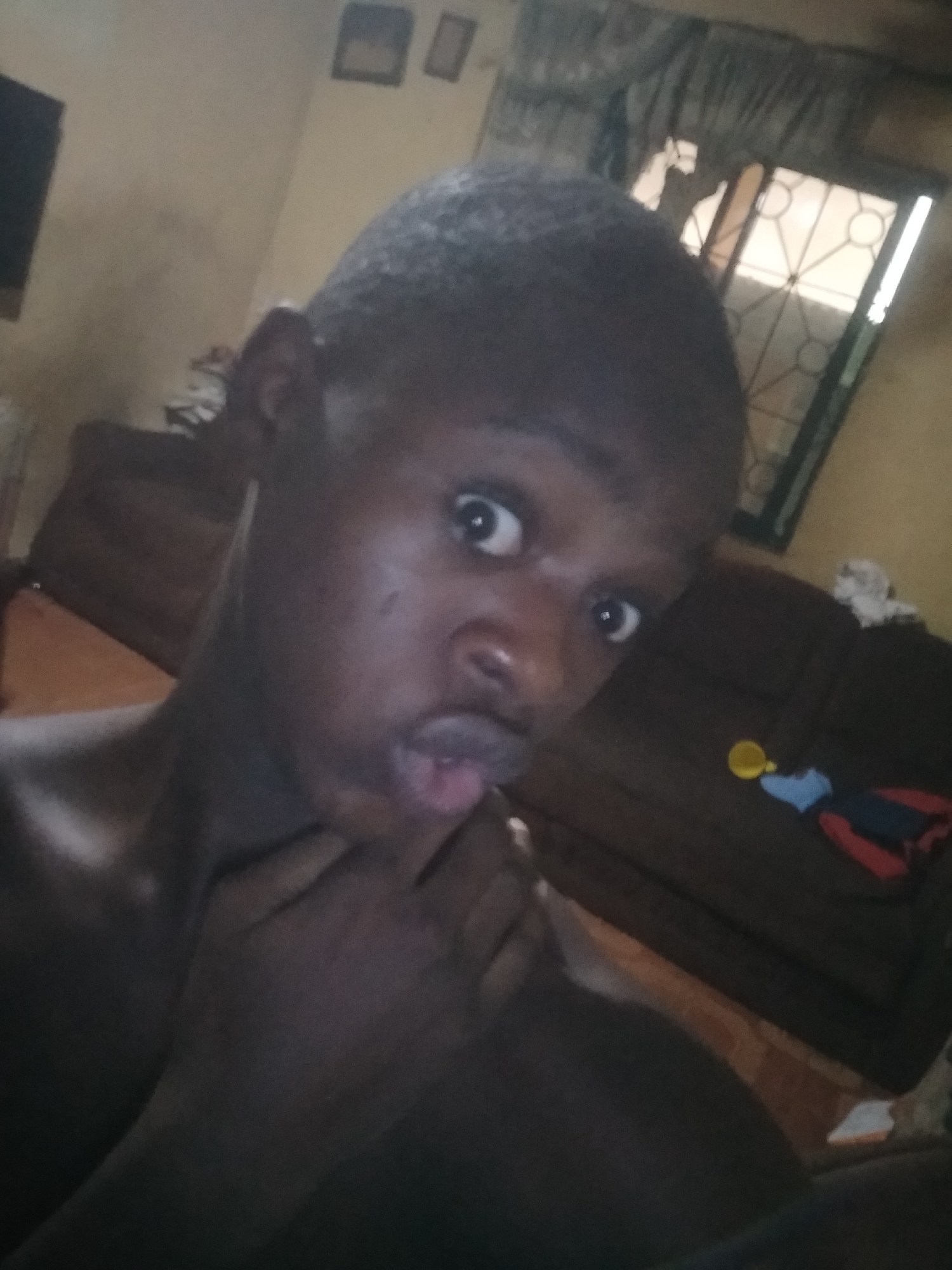 A selfie of a young black man with close-cropped hair is presented. He's indoors, likely in a residence in Shangisha, Nigeria, based on the surroundings. His hand is up by his mouth, and he has a surprised expression. The background includes a brown sofa, a window with a patterned metal frame, and framed pictures on the wall. A television is partially visible. There is a baby toy on the couch in the background.