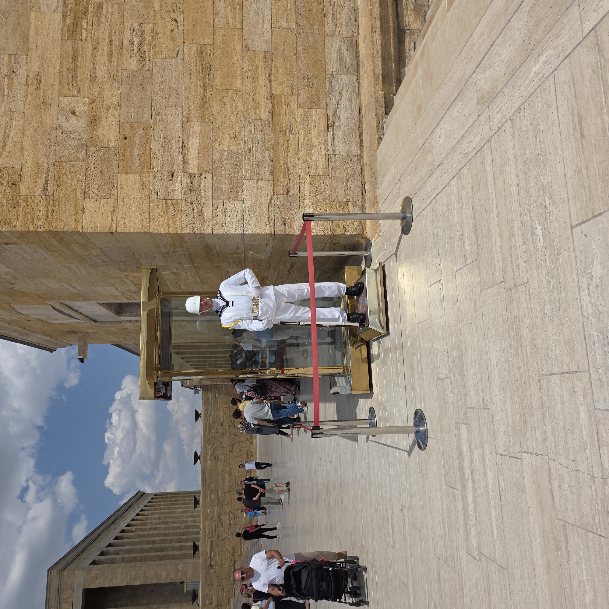 This is a photograph taken outdoors in Ankara, Turkey, likely at Anıtkabir, the mausoleum of Mustafa Kemal Atatürk. The central subject is a guard in a white uniform and helmet, standing at attention within a golden-trimmed glass enclosure. A red rope barrier with silver stanchions encircles the enclosure. In the background, the monumental stone architecture of a building, characteristic of Anıtkabir, is visible, with a stone relief frieze running along its base. Several people are walking on the light-colored paved ground in the middle and background of the image. One man in the foreground on the left is pushing a black baby stroller. The sky is blue with white clouds, indicating daytime.