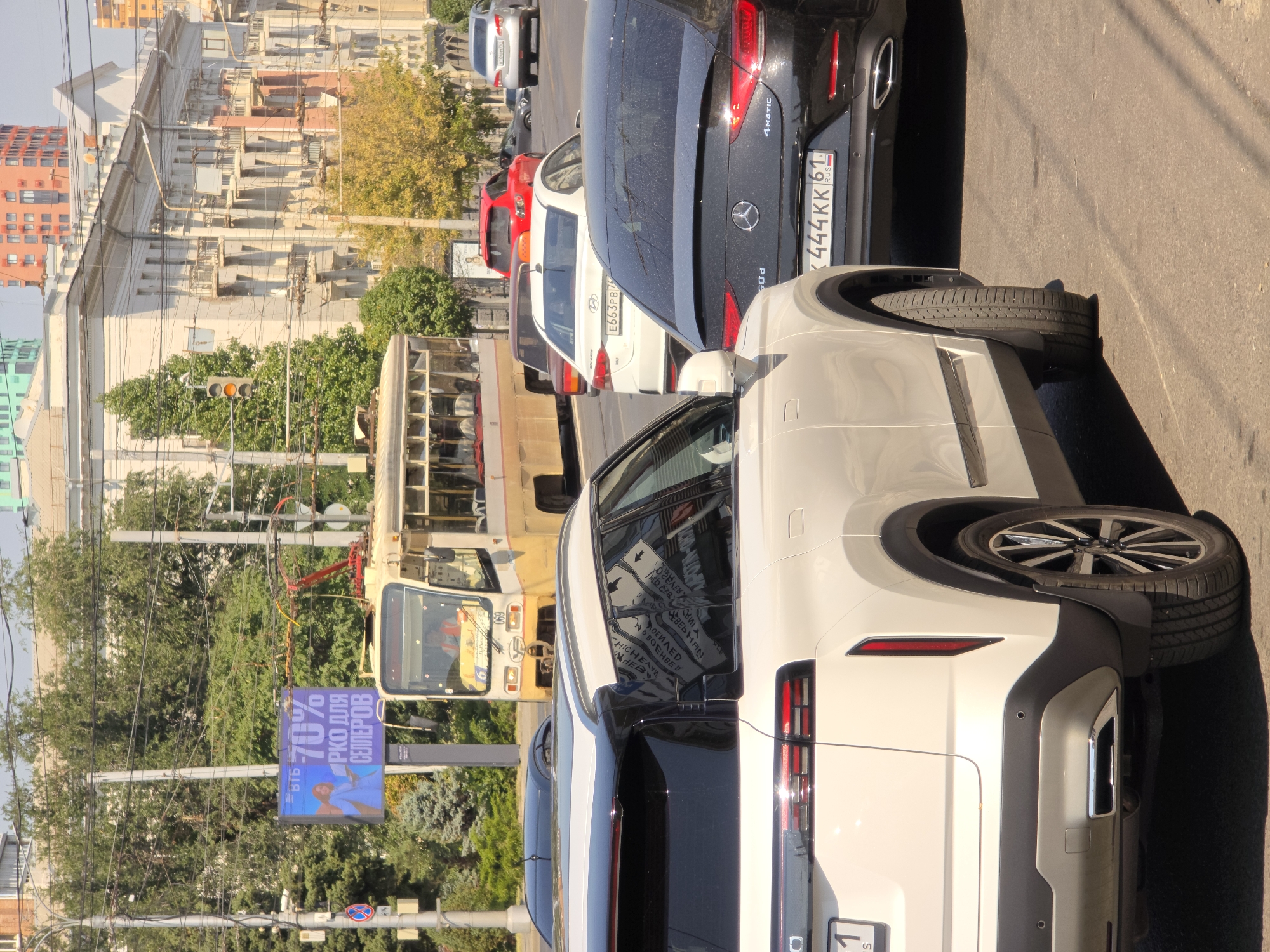 In Rostov-on-Don, Russia, a sunny daytime scene depicts a busy urban street with multiple vehicles in traffic. A white SUV is prominently positioned in the foreground, followed by a black Mercedes-Benz sedan. In the middle ground, a yellow and beige trolleybus, identified by the number 069, is visible, along with several other cars, including a red sedan and a white sedan. The background features ornate buildings with balconies and trees, suggesting an established city center. An electronic billboard displays the text "70%" and Russian text, indicating it is an advertisement. The overall impression is that of a typical city intersection during daylight hours.