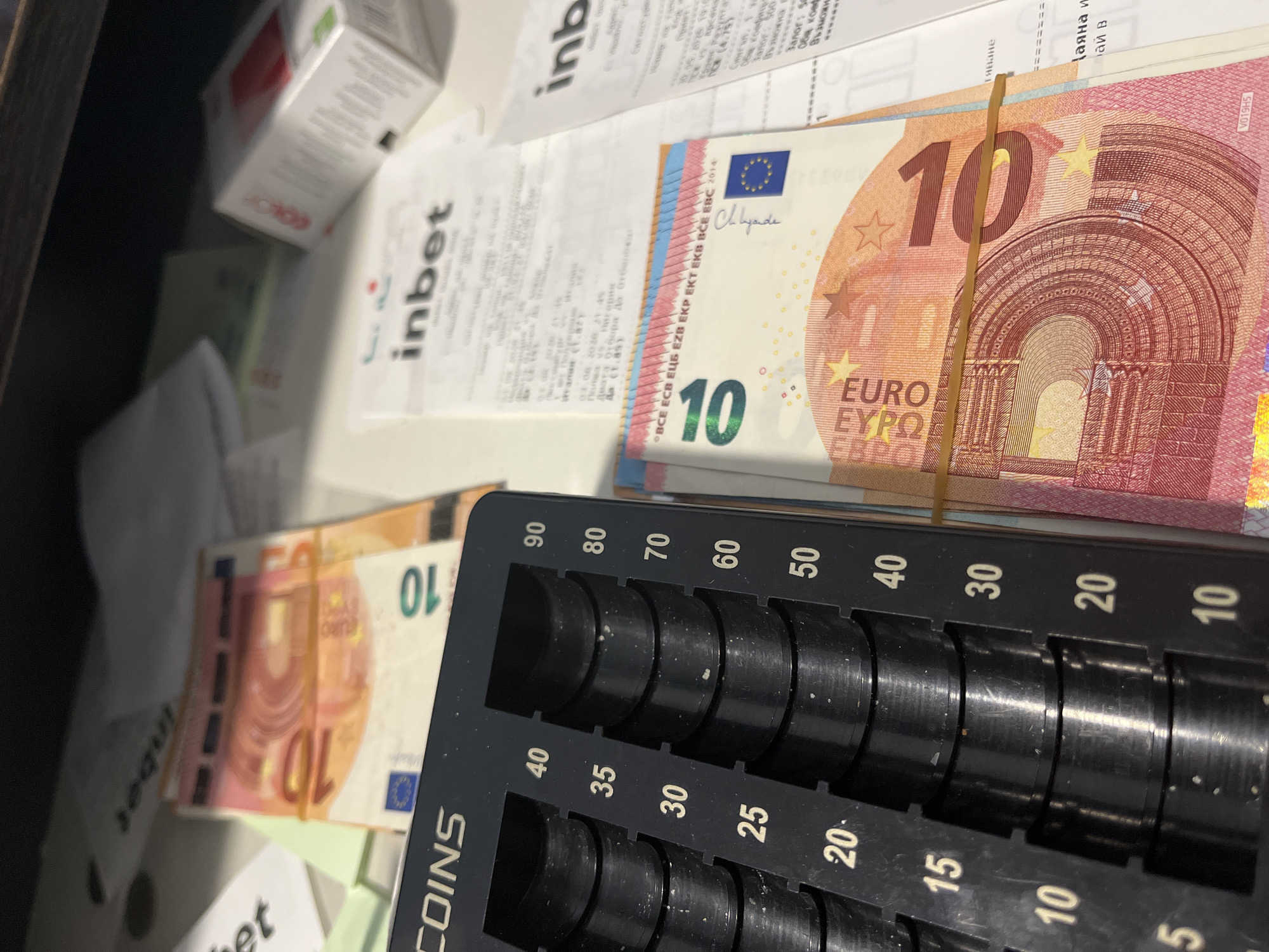 The image captures an indoor scene, likely a drawer or counter in a commercial setting in Ivailo, Bulgaria, where money handling takes place. The main subjects are two bundles of 10 Euro banknotes, secured with rubber bands, and a black coin counter.

One bundle of Euro notes is prominently displayed in the upper right, while another is partially visible on the left. The 10 Euro notes feature the "EURO EBPO ΕΥΡΩ" inscription and the signature of "C. Luyde" (Mario Draghi), with "BCE ECB ЕЦБ EZB EKP EKT EKB BCE EBC 2014" also visible.

A black plastic coin counter or sorter occupies the bottom right foreground. It has multiple circular slots for different coin denominations, marked with numbers such as 90, 80, 70, 60, 50, 40, 35, 30, 25, 20, 15, 10, and 5. The word "COINS" is visible on its side. The slots appear empty, suggesting it might be ready for use or recently used.

In the background and scattered around are several crumpled and flat paper receipts. Multiple receipts clearly show the logo and text "inbet," indicating a connection to a betting or gaming establishment. One receipt displays Bulgarian text and a date "01.06.2016 21:45," along with phrases like "Двата отбора да отбележа" (both teams to score), "Възможна" (possible), and "Общ коеф." (total odds), further supporting a betting context. A small white box, partially visible, has the word "COLOR" printed on it. The overall impression is of a busy cash handling area in a business that processes financial transactions and issues receipts, likely related to gaming or services in Bulgaria.