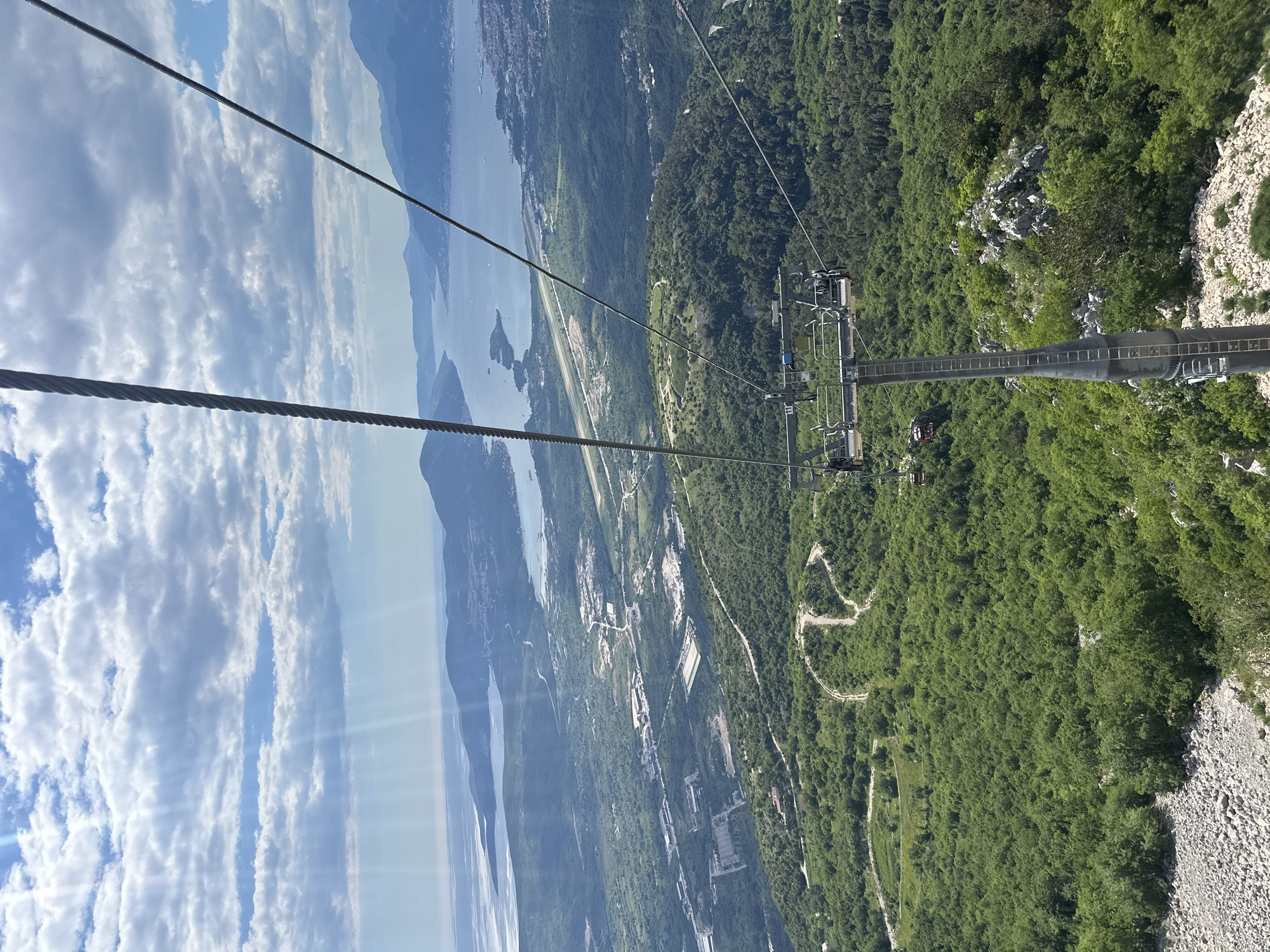 The image shows a view from a cable car ascending Lovćen Mountain near Čavori, Montenegro. The Adriatic Sea, the city of Tivat, and mountains form the backdrop. Dense forest covers the mountainside below, marked by winding trails. The cable car infrastructure, including supporting structures and cables, dominates the foreground. The sky has white clouds and a sunny sky.