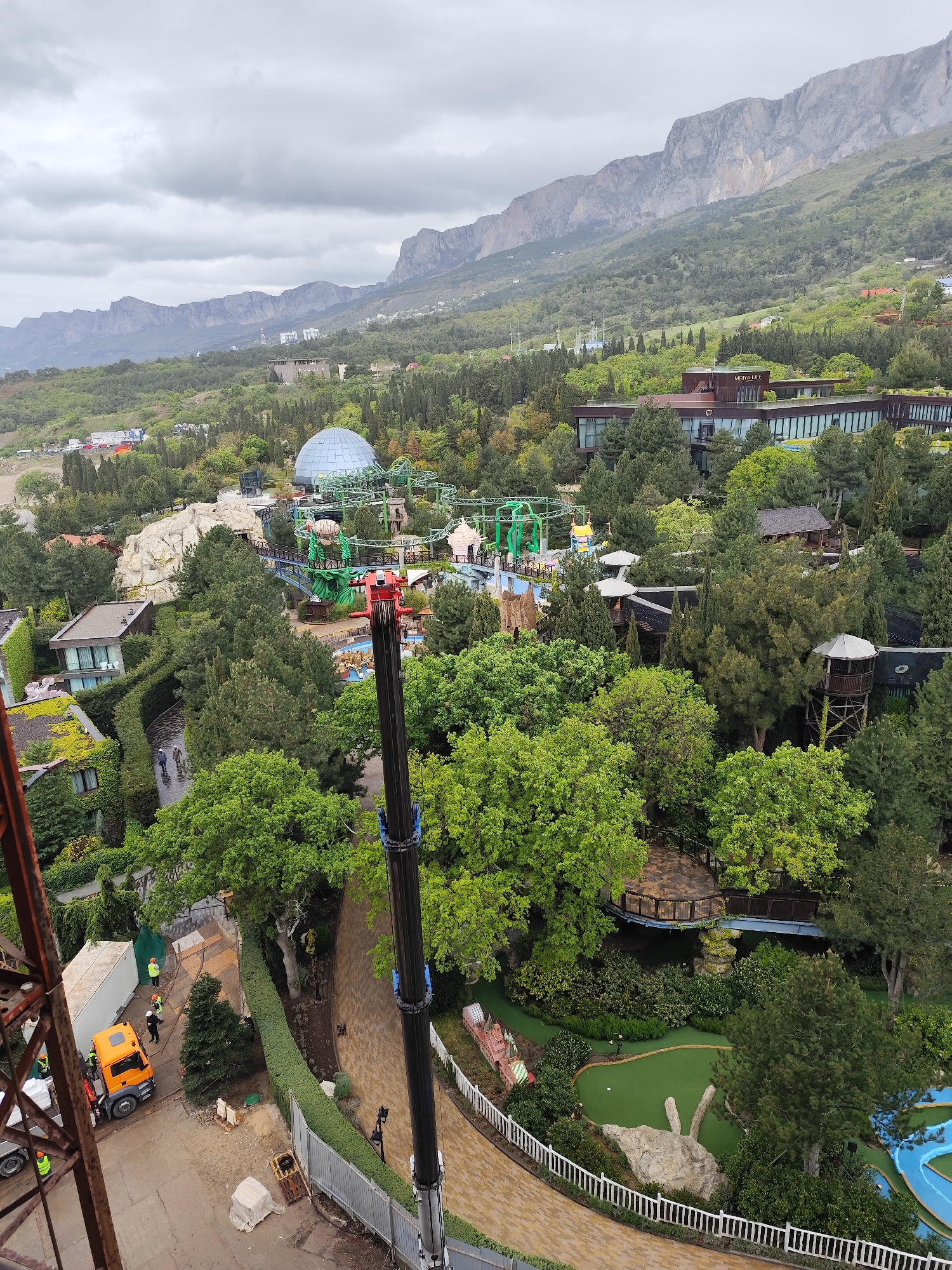 The image shows an aerial view of a landscape in Симеизский поселковый совет, Ukraine. In the distance are mountains under a cloudy sky. In the foreground is a construction scene, with a crane and workers near a truck. There is a walkway along a green lawn with a white fence. Between the foreground and the distant mountains are features including a dome, a rollercoaster, buildings, and trees.