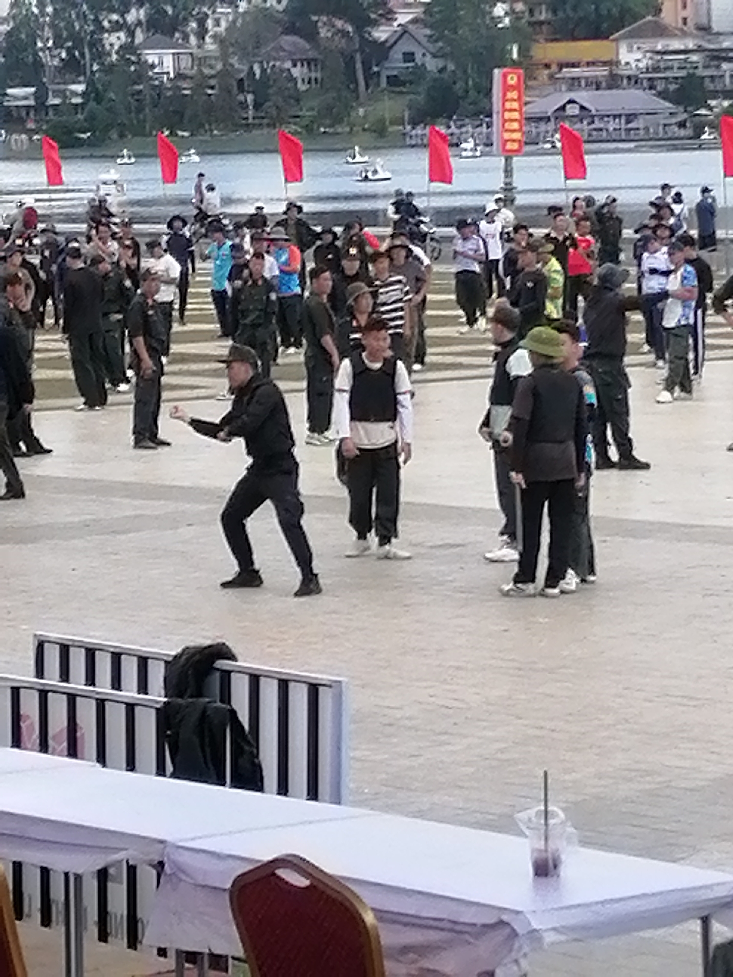 The image depicts a wide outdoor plaza in Da Lat, Vietnam, with a body of water in the background, likely Xuan Huong Lake given the context. A group of people, many of whom appear to be male, are gathered in the plaza, dressed in casual attire, with some wearing vests. Several individuals are engaged in what seems to be a coordinated movement or exercise. In the foreground, two tables are set up with white tablecloths. Red flags are visible in the background, as well as buildings suggesting a town or city setting. The overall scene appears to be a public event or gathering taking place in Da Lat.