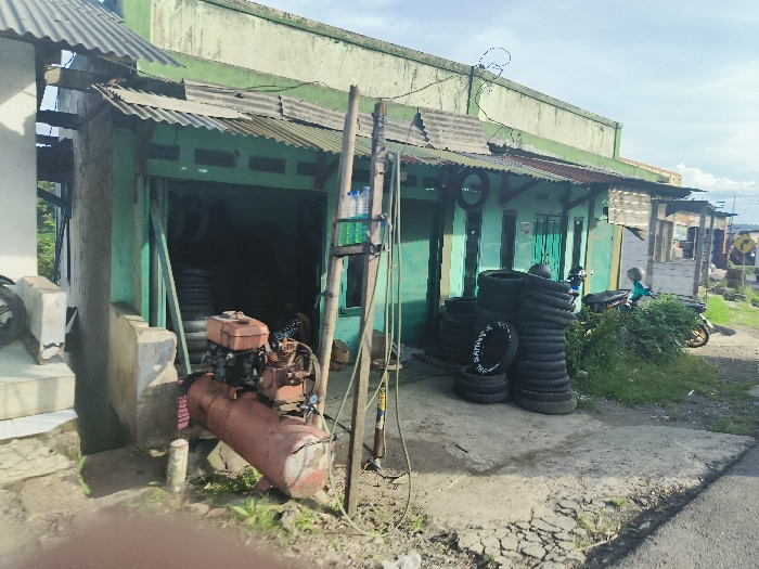 The image shows a small, green, corrugated metal building in Cikalong, Indonesia. The building appears to be a tire shop. A stack of tires is leaning against the wall in front of the entrance to the shop. To the left of the tires is a red, gasoline-powered air compressor. In front of the compressor, the ground is covered in grass. To the right of the tires, there is a green motorcycle. The motorcycle has a black helmet on the seat. The background shows more green corrugated metal buildings and green foliage. The building appears to be on the side of a dirt road. The coordinates provided, -6.731, 107.444, are likely in the city of Cikalong, Indonesia. It is impossible to tell the specific location with the provided coordinates. There are no people visible in the image.