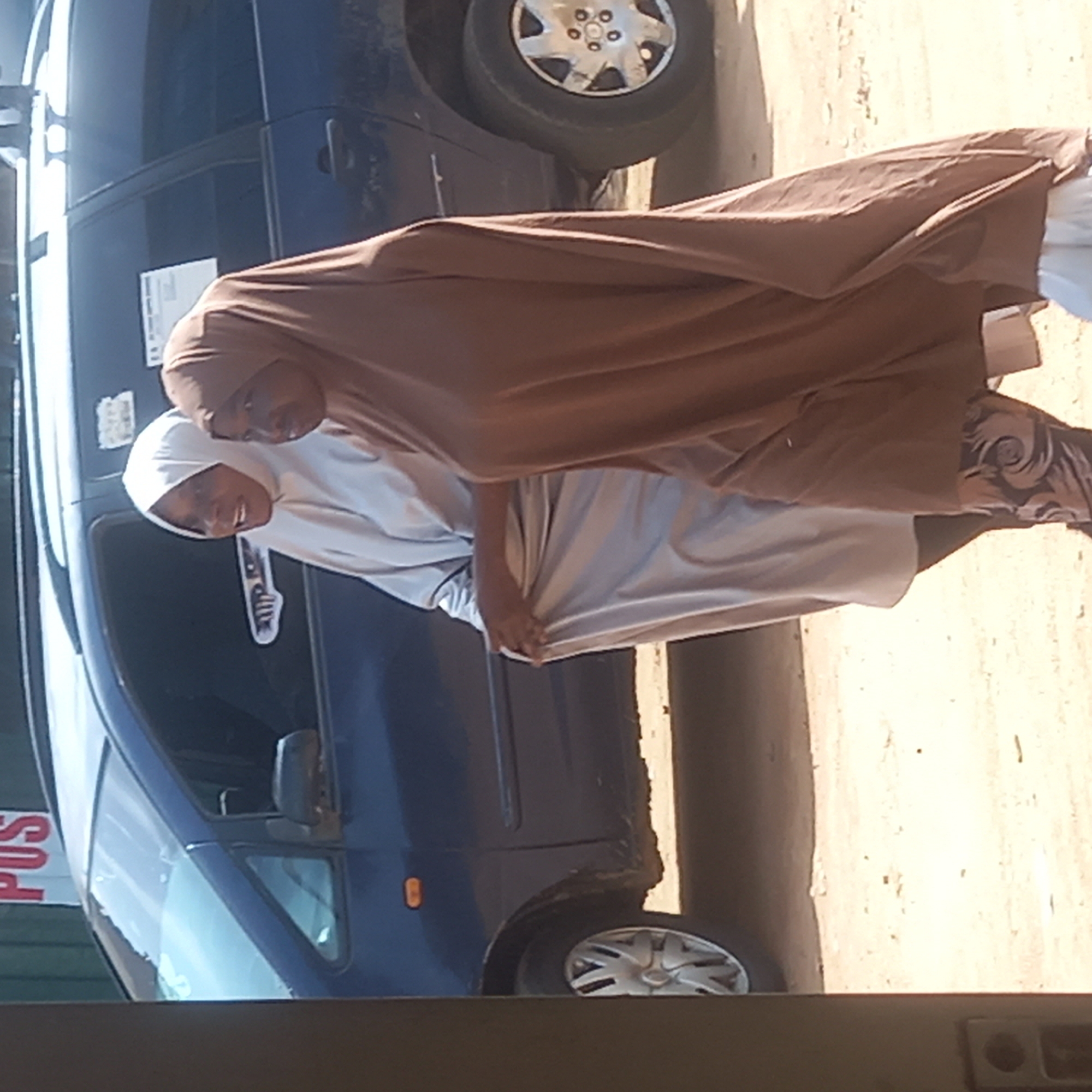 Two women stand together in Sardauna, Nigeria, next to a dark blue minivan. The woman on the right wears a brown hijab and patterned pants. The woman on the left wears a white hijab and a light gray garment. They are holding hands and smiling. The minivan is parked on a street next to a beige-colored wall. "POS" is visible on a sign near the minivan. The ground is tan and appears to be dirt or sand.
