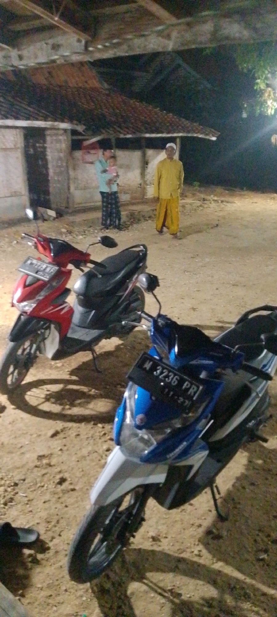 In Pangereman, Indonesia, at night, two scooters are parked on dirt ground. One scooter is red and black, and the other is blue and white with a license plate reading "M 3236 PR". In the background, a man in a yellow robe and white cap stands, and another person in a blue shirt and patterned pants holds a baby. A simple building with a tiled roof is visible behind them. The scene appears to be an outdoor gathering.