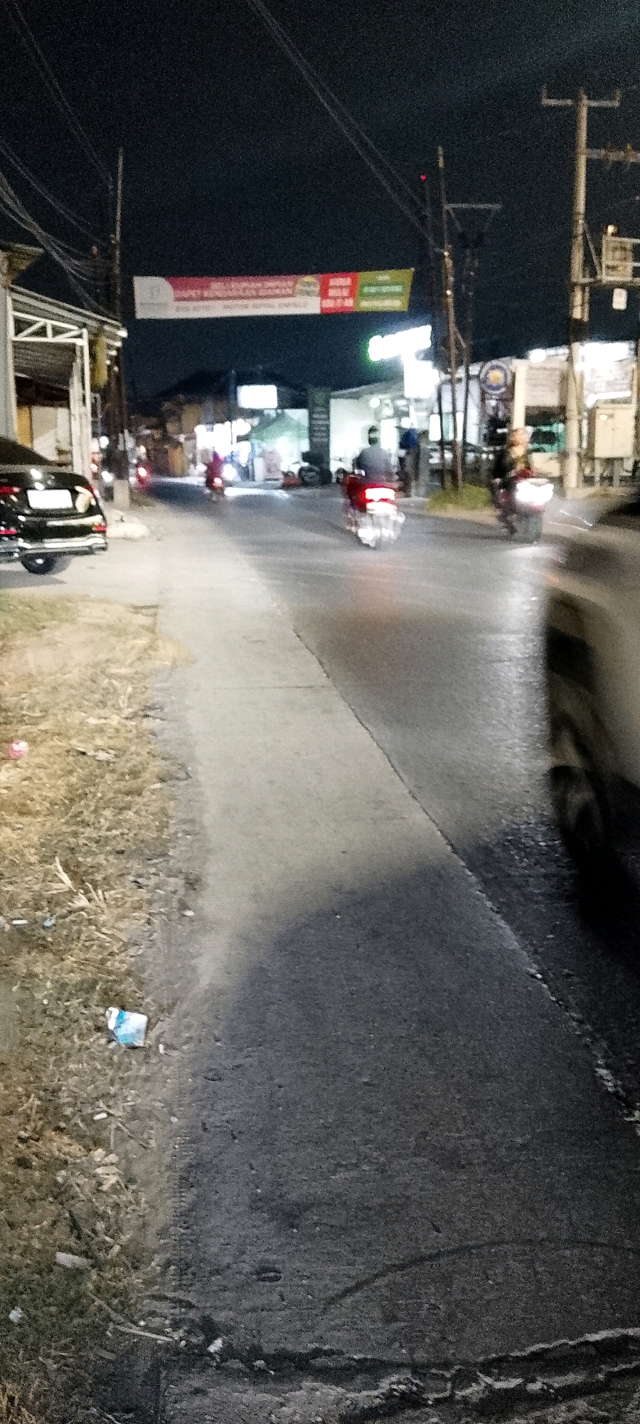The image depicts a street scene at night in Satriamekar, Indonesia. A dark-colored car is parked on the left side of the road. Several motorcycles are visible on the street, with two in motion. One motorcycle in the foreground, on the right, is blurred due to motion and its headlight is illuminated. Another motorcycle, with a rider wearing a dark helmet and jacket, is moving away from the camera in the center of the road, its taillight illuminated. A third motorcycle, further in the background, also has its taillight on. A banner with red and green sections is suspended across the street overhead. The environment includes a sidewalk or pathway on the left, bordering a patch of dry grass and some debris. Streetlights and overhead wires are present. The overall scene suggests a typical night view of a street in a developed area.