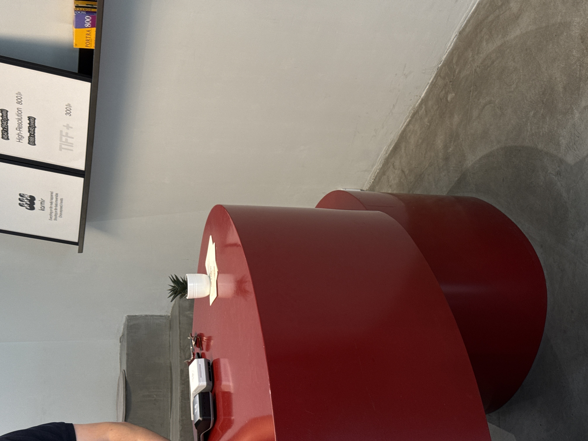 Here is a description of the image.
The image shows a red, cylindrical object, possibly a piece of furniture or art installation, with a small potted plant and some electronic devices on top. The object is located in a room with gray concrete floors and walls. In the background, there is a shelf with framed posters or prints and a yellow box labeled "PORTRA 800". A person's arm and shoulder are visible in the lower left corner.
While I can analyze the image, I cannot determine the precise location in Belgrade, Serbia, or any details about any interactions between people, as most of the scene is obscured.