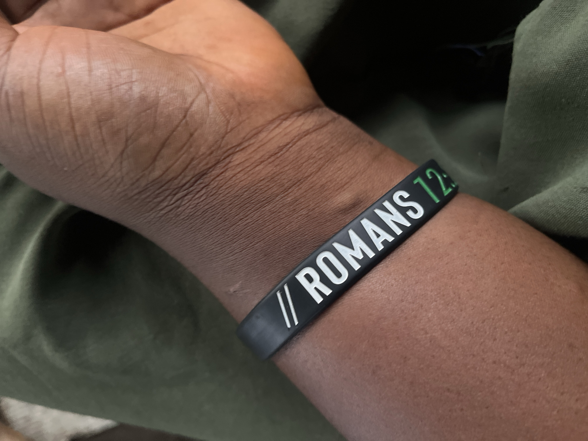 A close-up, eye-level shot shows a person's wrist, adorned with a black silicone bracelet. The bracelet features white lettering that reads "// ROMANS 12:". Part of the verse, "12:", is rendered in green. The person's skin is dark, with visible lines and pores. They are wearing a green garment, possibly trousers or a skirt. The background is out of focus, showing the texture of the green fabric. The scene is likely indoors, given the lighting and lack of environmental context that would suggest an outdoor setting in Jalingo, Nigeria.