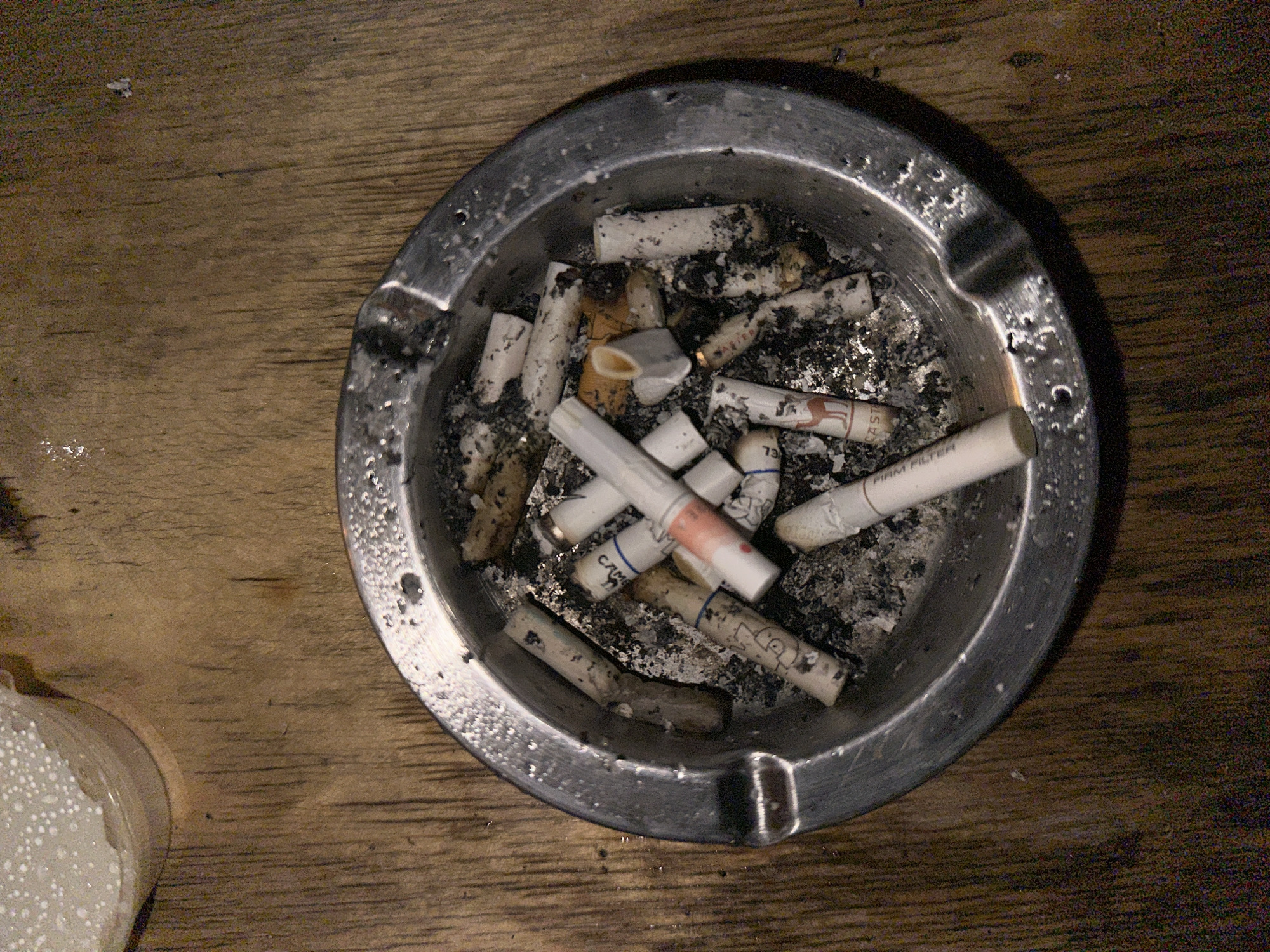The image depicts a silver ashtray, filled with numerous extinguished cigarette butts and ash. The ashtray rests on a wooden surface. A blurry object, possibly a container, is visible in the lower left corner of the image. The location is unknown.