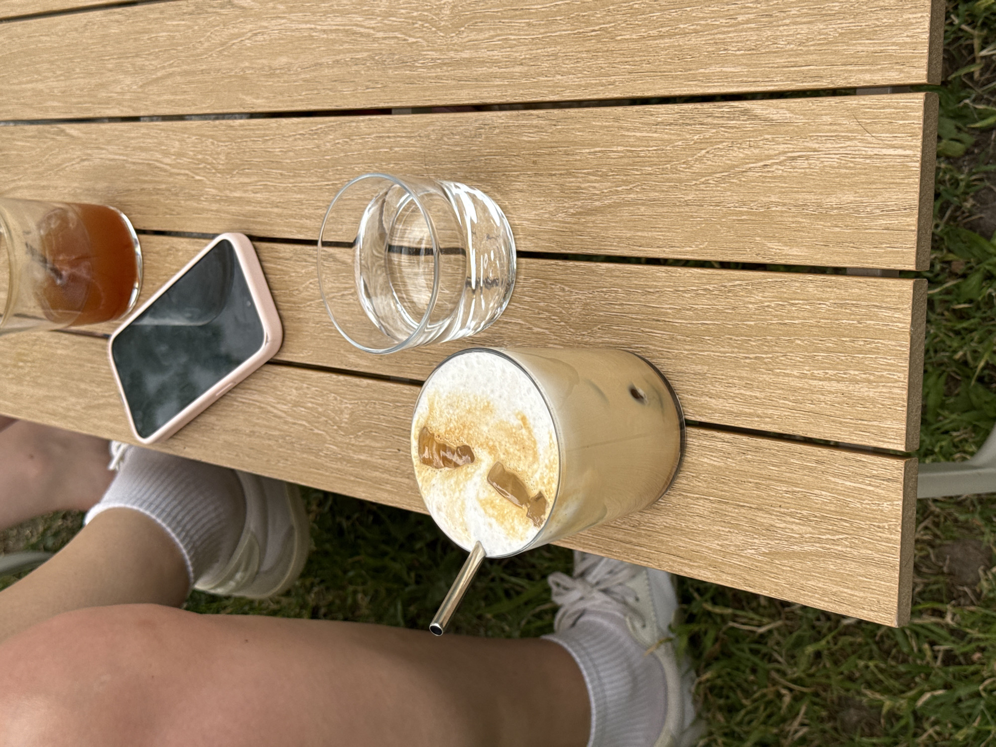 A top-down shot shows a wooden table with several items and part of a person sitting at it. The table is made of light brown wooden slats. On the table are, from left to right: a glass of amber liquid, a smartphone with a pink case and a black screen, an empty clear glass turned upside down, and a glass of iced coffee with a silver straw. The iced coffee has a foamy top with some ice cubes on it. Under the table, a person is sitting with their legs visible, wearing white socks and sneakers. The background shows some green grass. The city of Belgrade, Serbia is not confirmed from visual elements.