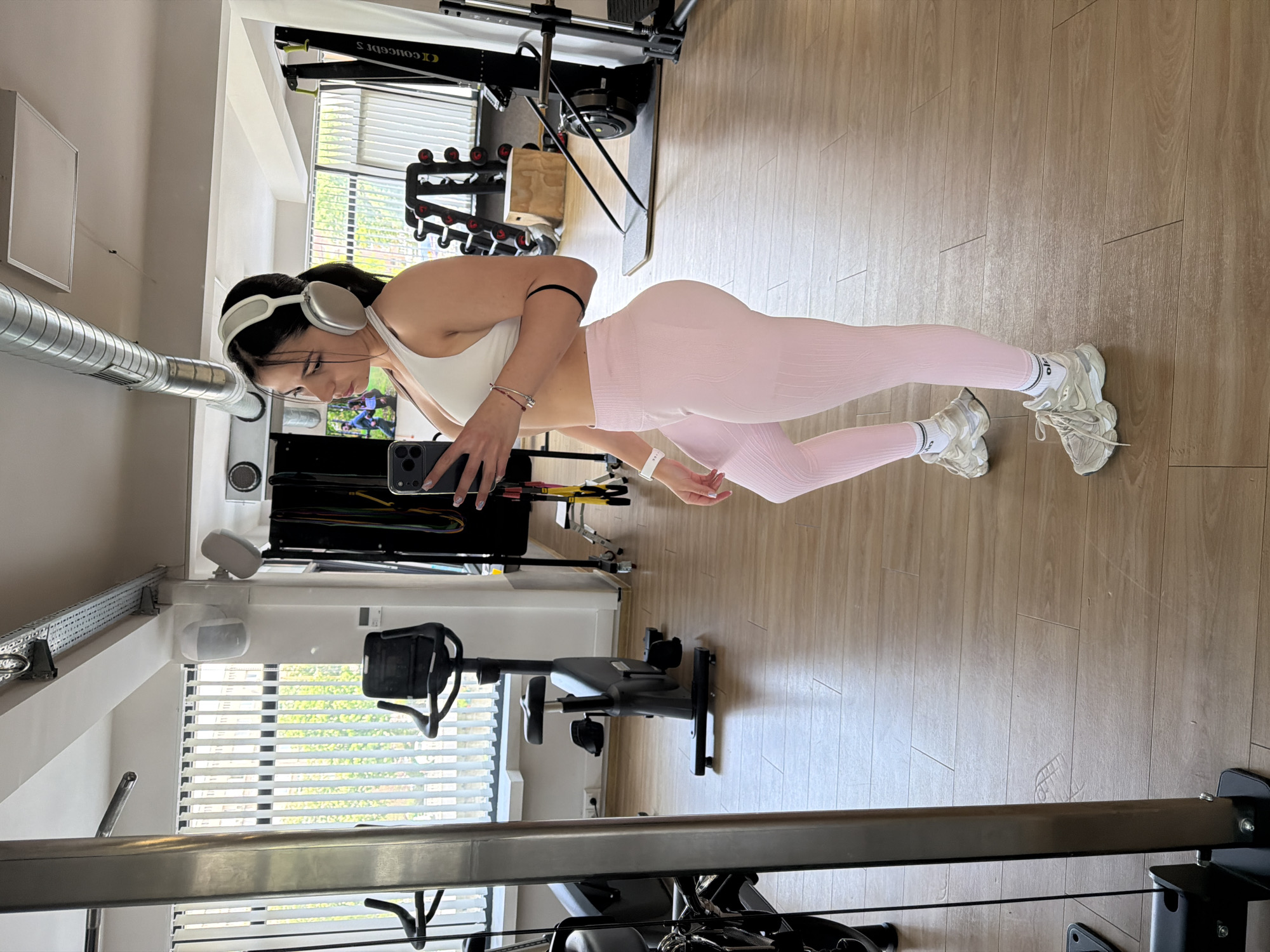 A woman in a gym is taking a selfie. She is wearing a white sports bra, pink leggings, and white sneakers with white socks. She also has white headphones on and a black band on her arm. She is holding a smartphone in her left hand. The gym has a rowing machine, weights, and a stationary bike. The floor is made of wood. There is a window in the background. The city of Gabrovo, Bulgaria, is not discernible in the image.
