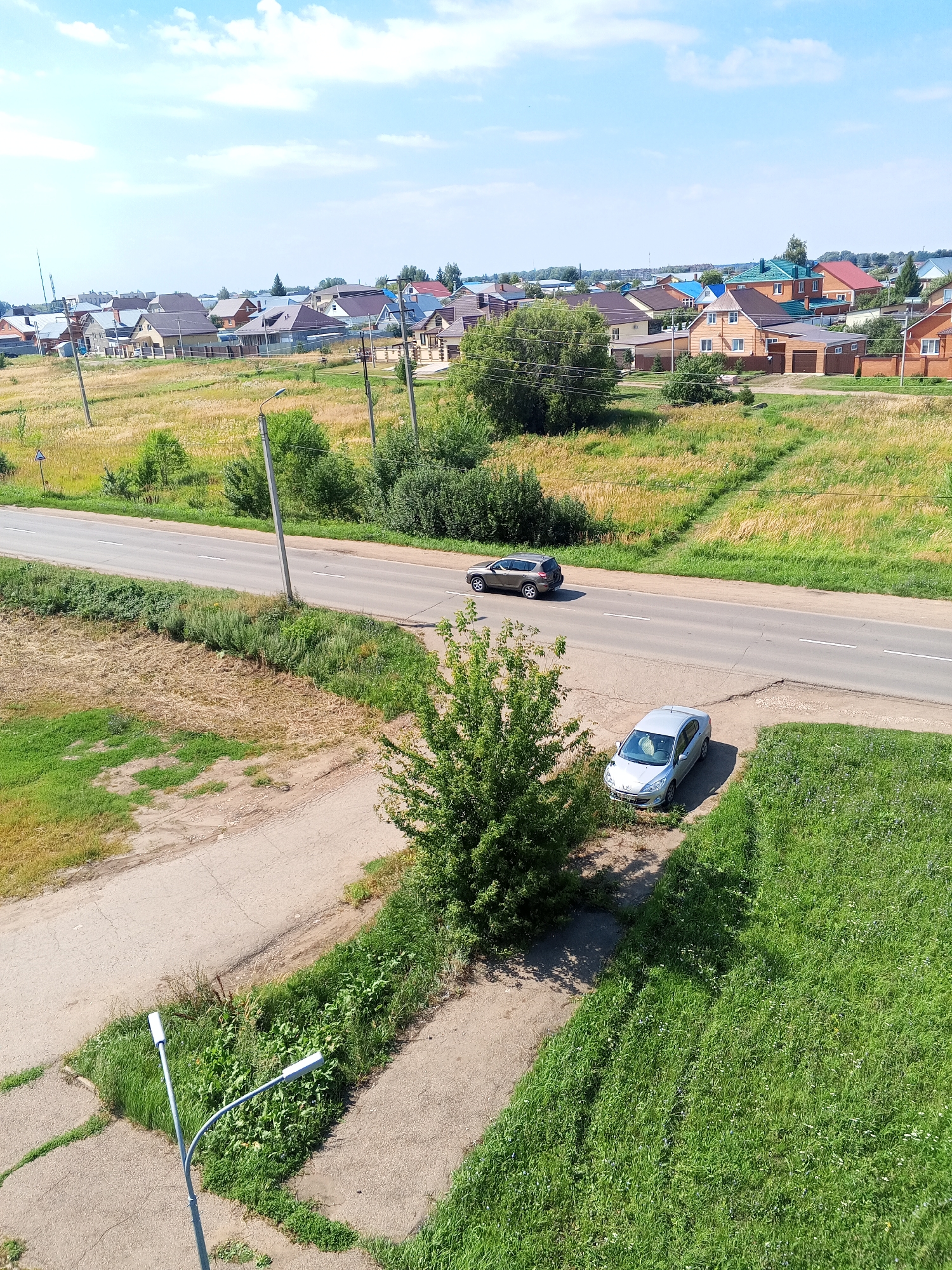 The image shows a rural or suburban residential area with a road running through it. Several houses with varied roof colors are visible in the background, suggesting a developed community. Two cars are present on the road: a dark SUV in the distance and a silver sedan parked on the side of the road closer to the foreground. The environment is characterized by green grass, some trees, and a dry, grassy field. The sky is blue with scattered white clouds, indicating fair weather. The presence of utility poles and streetlights suggests infrastructure common to populated areas. The overall scene depicts a tranquil day in a residential neighborhood, possibly in Chistopol, Russia, given the context.