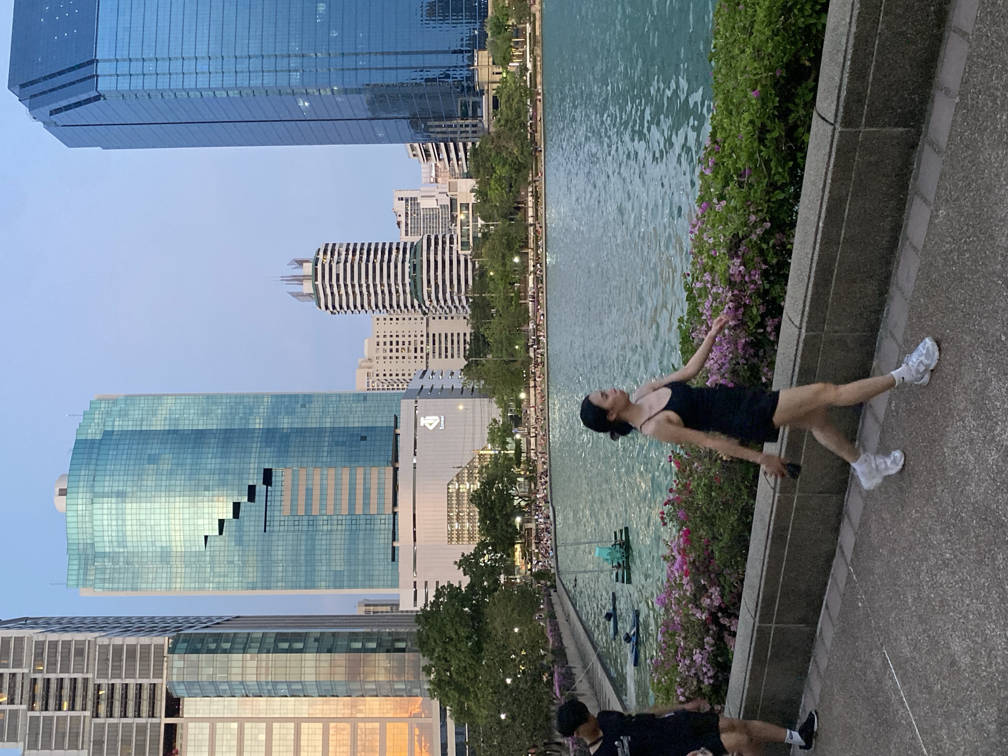 The photo shows a person in a black dress and white sneakers walking along a promenade in Lumphini Park, Bangkok, Thailand. The park borders a large pond filled with water, and the far side is lined with people. Lush greenery and vibrant purple flowers grow along the promenade's edge. Skyscrapers dominate the background. The sky is a gradient of blue and hints of orange. There is another person wearing black on the lower left.