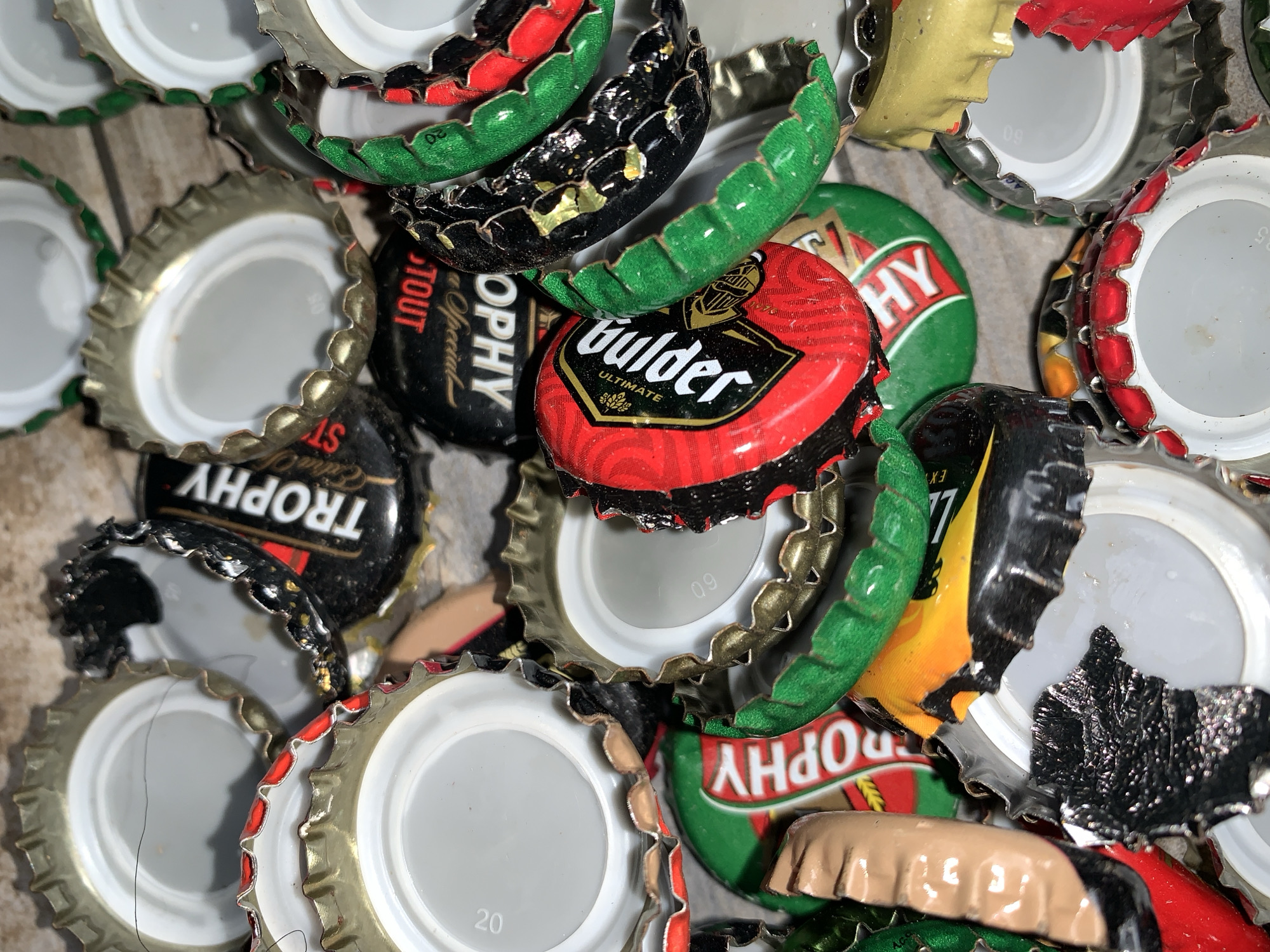 A collection of approximately two dozen metallic bottle caps is depicted, observed in New Stadium, Nigeria. The caps exhibit varying degrees of deformation, including bending, crumpling, and flattening. Predominant colors among the caps include red, green, black, and gold, with most interior liners being white or light grey. Visible branding includes "TROPHY" in white lettering on several caps with black or green backgrounds, "Gulder ULTIMATE" on a red and black cap, and the word "STOUT" on an inverted black cap. Numerical identifiers such as "20" and "09" are visible on the interior surfaces of some liners. A fine, dark strand is present across the white interior of two caps in the upper left portion of the image. The bottle caps are resting on an out-of-focus, light-colored, textured surface. No people, actions, or specific environmental features beyond the immediate surface are discernible.