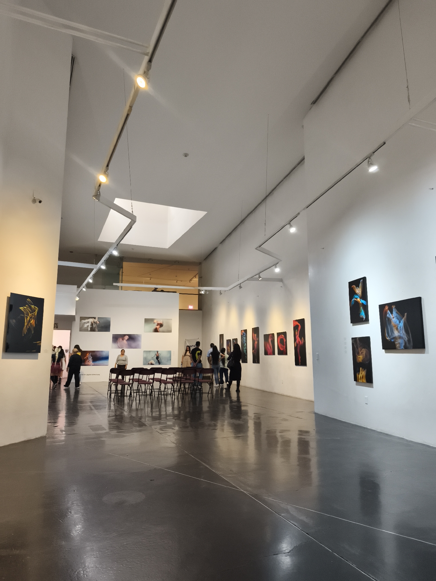 The image captures an interior view of an art gallery, likely located in the city of Querétaro, Mexico. The space is characterized by a high ceiling, a gray polished concrete floor, and white walls, typical of modern gallery design. A series of artworks are displayed along the walls, illuminated by track lighting. In the foreground, on the left, there is a black and gold abstract painting. Further down the gallery, on the left wall, there are multiple photographs. On the right wall, groupings of three abstract paintings are visible.
Several people are present in the scene. Near the center of the gallery, a group appears to be gathered around a row of maroon chairs lined up. Some people are looking at artwork, while others seem to be conversing. The attire of individuals suggests a casual and relaxed atmosphere.
The architecture includes a rectangular skylight in the ceiling, adding natural light to the gallery. The overall environment is well-lit and spacious, conducive to viewing art. The chairs suggest the space may be used for presentations.