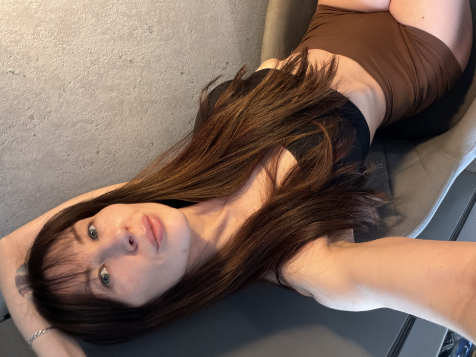 The image displays a woman reclining indoors, likely on a dark upholstered chair or sofa, positioned against a light gray, textured wall. The specific location within Perm, Russia, is not identifiable from the visual content. The woman, appearing to be in her 20s or 30s, has long, dark brown hair with reddish-brown highlights and bangs. She possesses blue-green eyes and full lips. Her attire consists of a black bra or bralette and brown high-waisted shorts. Her right arm is raised and bent, with her hand positioned above her head; a silver-colored chain bracelet is visible on her right wrist. She is looking directly at the camera, indicating a posed photograph.