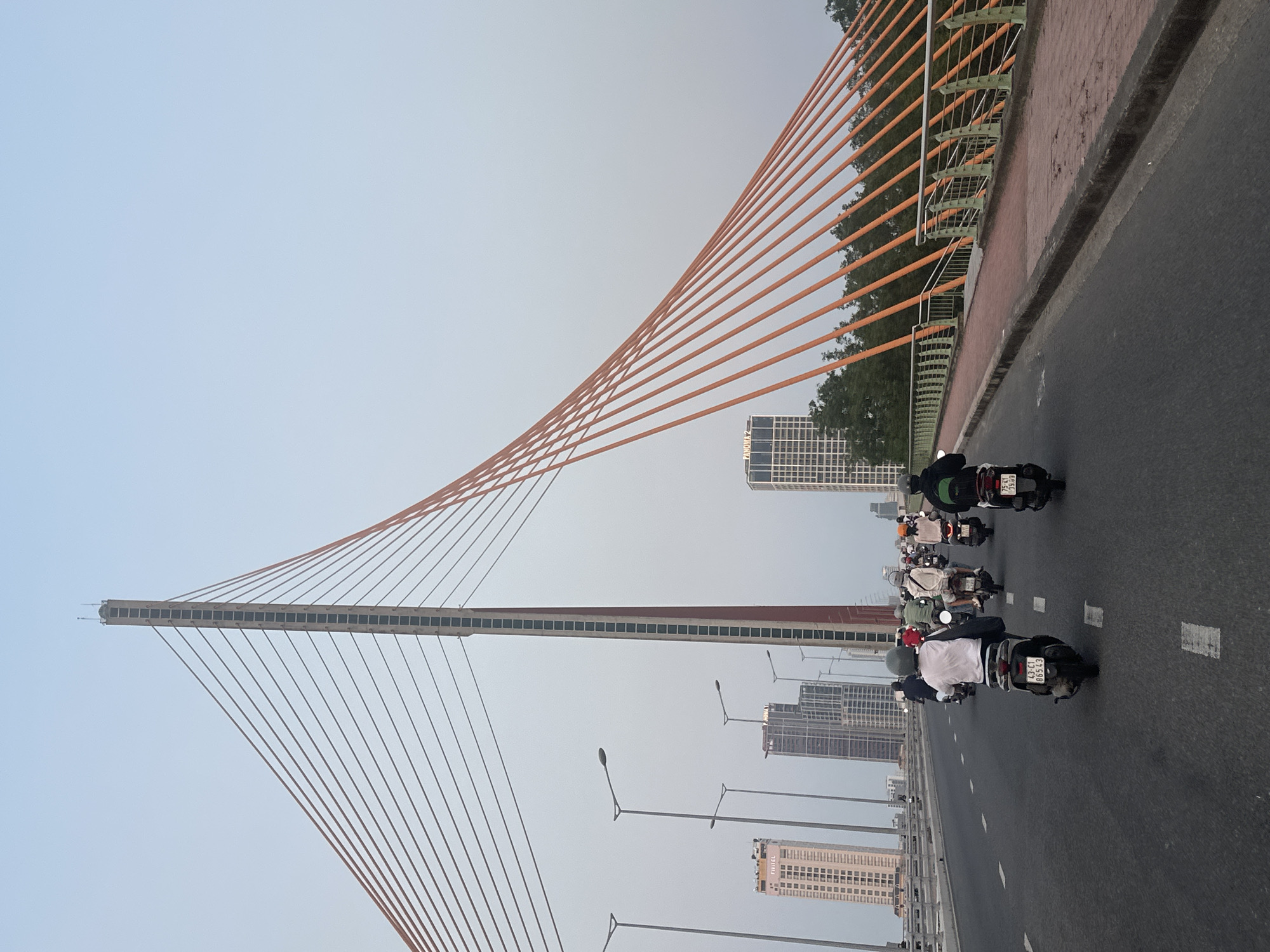 The image shows a view from a bridge in Đà Nẵng, Vietnam, with traffic moving along the road. The bridge is a cable-stayed type, notable for its orange support cables and a tall central tower. Several motorbikes are visible, representing the common form of transportation in the city. Buildings of varying heights, typical of a developing urban skyline, are present in the background. Streetlights line the road, indicating a modern infrastructure. The sky is overcast, creating a diffused lighting across the scene.