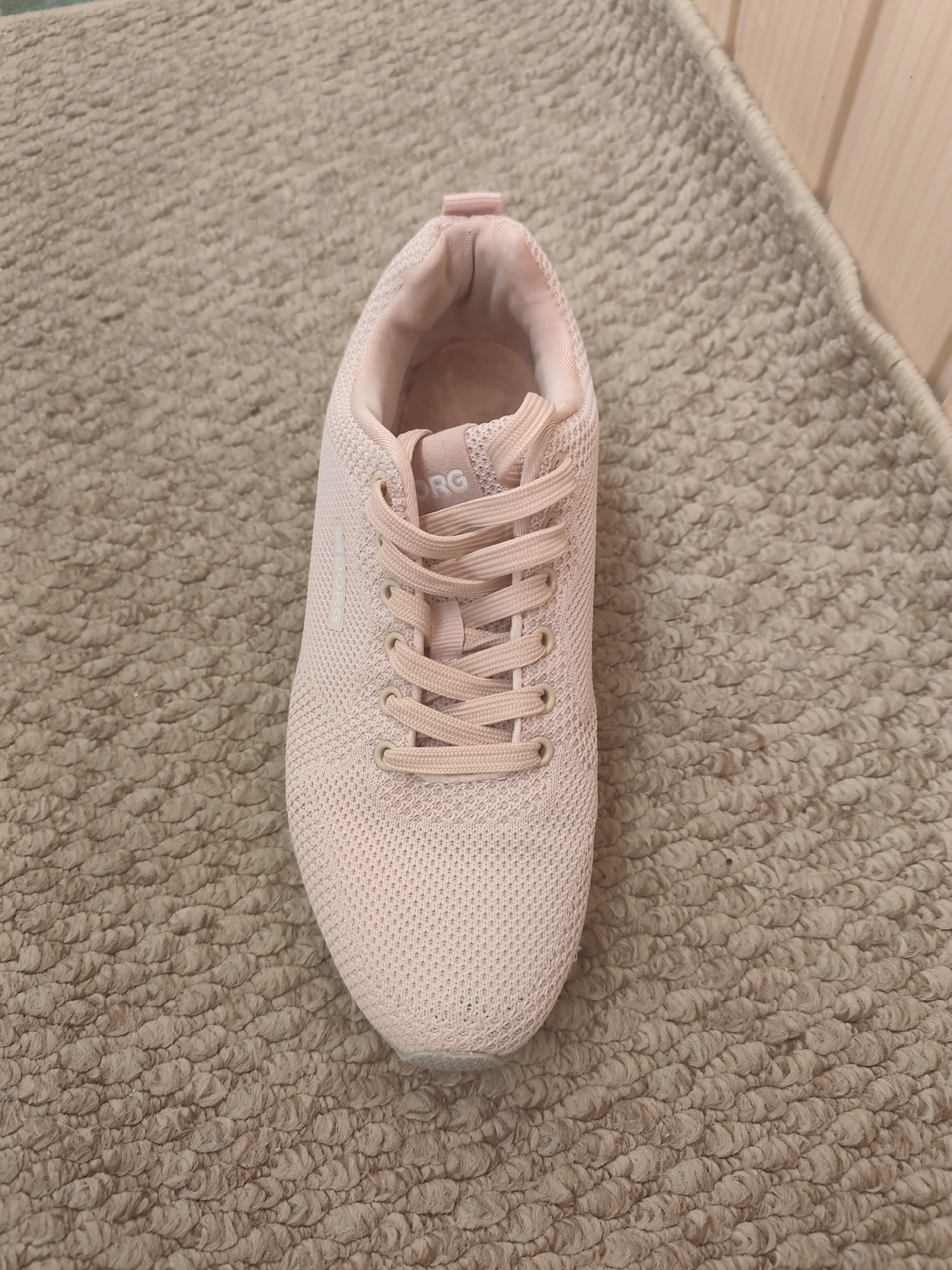 A single pink athletic shoe is lying on its side on a textured beige carpet. The shoe is made of a breathable mesh material and has light pink laces. The word "BORG" is visible in white lettering on the side of the shoe, and "ORG" is visible on the tongue. The background consists of the carpet and a light-colored wooden wall to the right. There is no indication of the shoe's location in the city of Kyiv, Ukraine, or any people or actions depicted in the image.