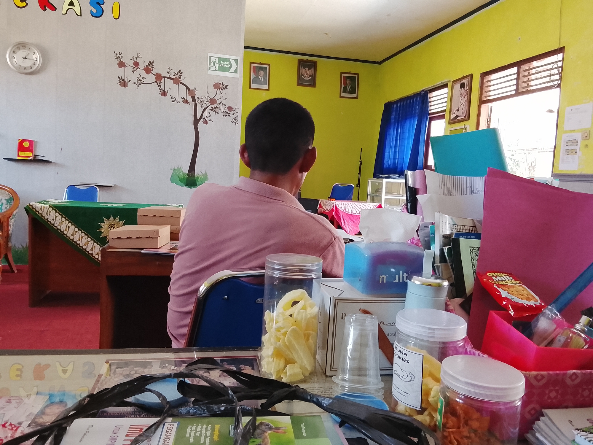 In the city of Kebasen, Indonesia, a man in a pink polo shirt is seated facing away from the camera, looking towards a desk. The room has bright yellow walls and a gray wall adorned with a decorative tree mural. On the gray wall, a circular clock hangs near a framed "JALUR EVAKUASI" (evacuation route) sign. Three framed portraits are displayed on the yellow wall. A large window with wooden blinds allows sunlight to enter the room. The desk in the foreground is covered with various items, including boxes, jars of snacks, and what appears to be paperwork. A blue chair is positioned behind the man, and a portion of a red carpet is visible. The overall impression is that of an office or classroom setting.