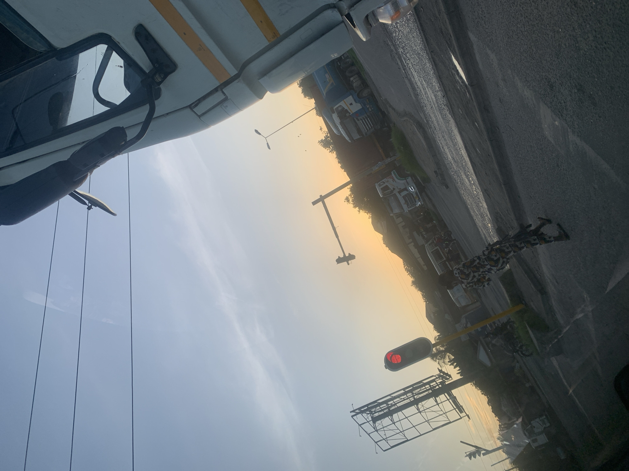 Captured from the high vantage point of a vehicle, the image shows a street scene in Dar es Salaam, Tanzania. A red traffic light signals a halt, mounted above an empty billboard frame. A line of trucks and other vehicles are visible along the roadside. A pedestrian is discernible near the edge of the road, dressed in clothing with a mixed pattern. The sky appears hazy, illuminated by the sun, suggesting either dawn or dusk. Power lines are visible against the light sky. The road is paved, and wet in places, hinting at recent rain. The foreground includes part of the vehicle from which the photo was taken, capturing a side mirror and part of the vehicle's structure.