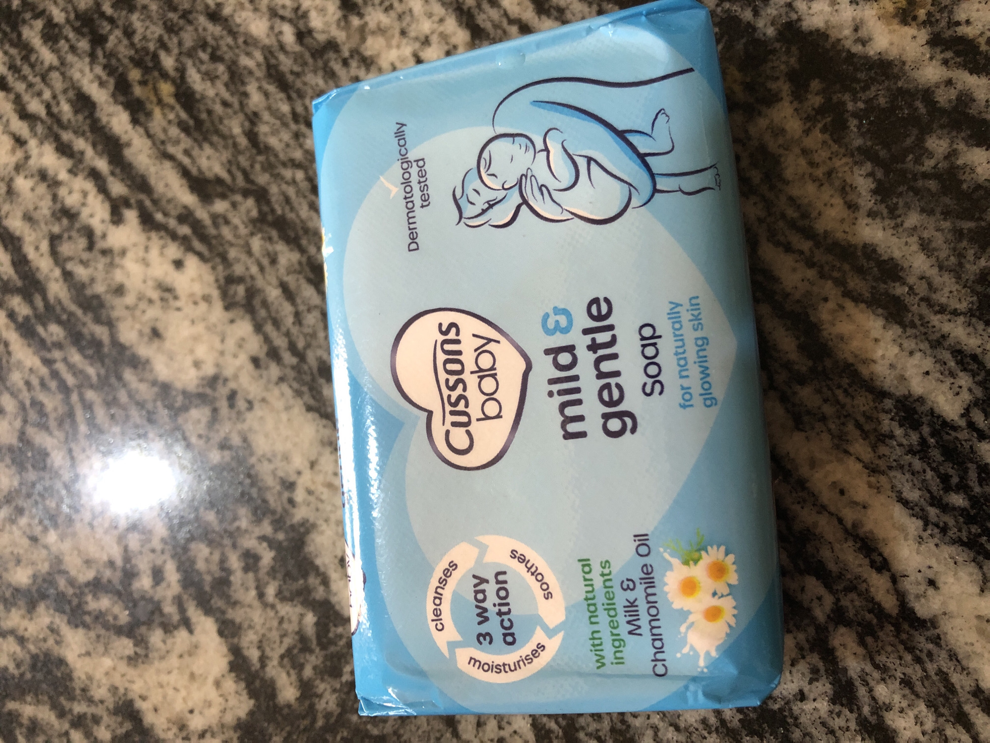 This is a media file of a Cussons Baby Mild & Gentle Soap bar. The soap bar is encased in a light blue wrapper featuring an illustration of an adult holding a baby. The text on the wrapper indicates that the soap is "Dermatologically tested" and provides a "3 way action" that "cleanses," "soothes," and "moisturises." It also states that the soap is made "with natural ingredients Milk & Chamomile Oil" for "naturally glowing skin." The soap bar is placed on a dark, speckled surface that appears to be a countertop or table. The lighting suggests it could be indoors, possibly during daylight hours. The overall impression is one of cleanliness and gentleness, aligning with the product's branding for baby care. There are no explicit clues about the specific time of day, weather, or other people present in the scene. The background is largely out of focus, directing attention to the product itself.
